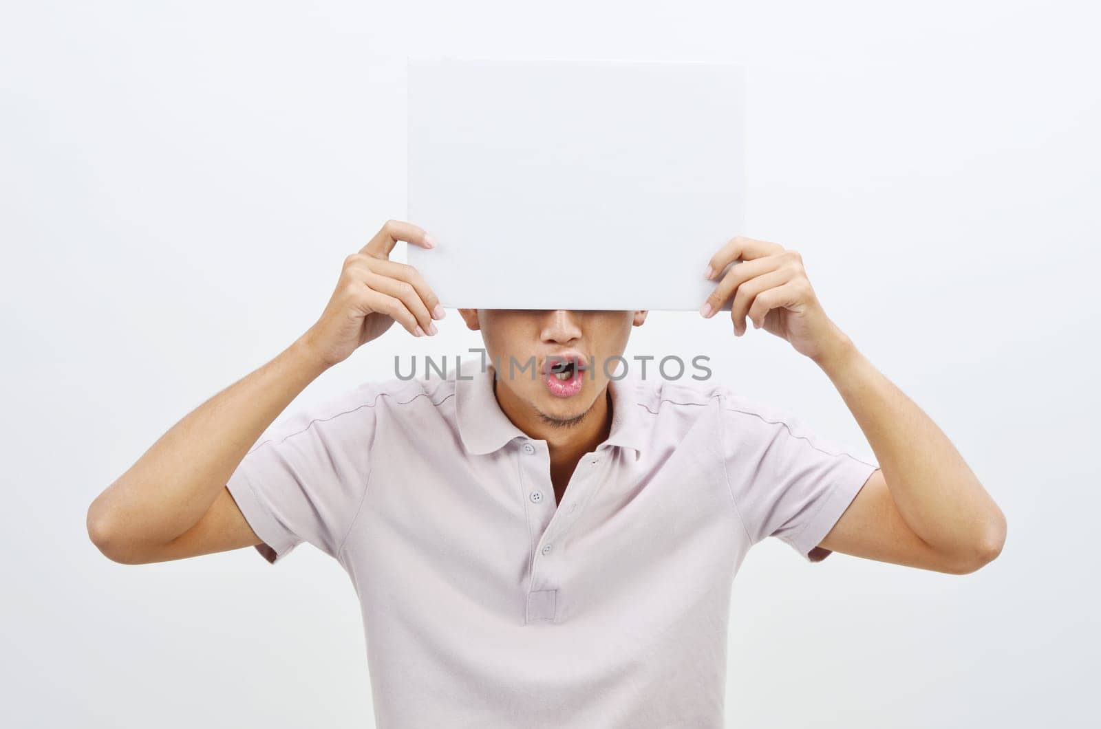 Portrait of shocked Asian man hand holding white blank paper card covering his eyes, standing isolated on plain background.