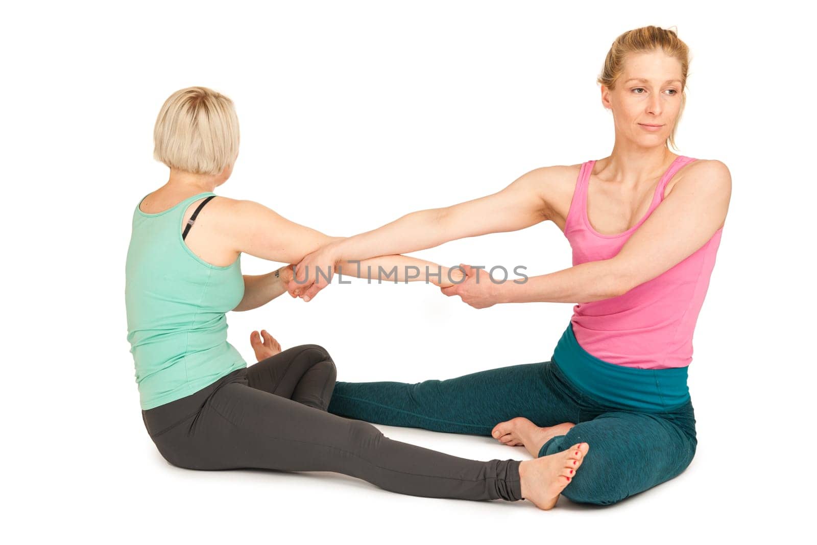 Side full-body view of two young blonde women sitting on the floor,touching each other on the soles of their feet and holding hands during a partner yoga exercise against a white background with slight shadows.