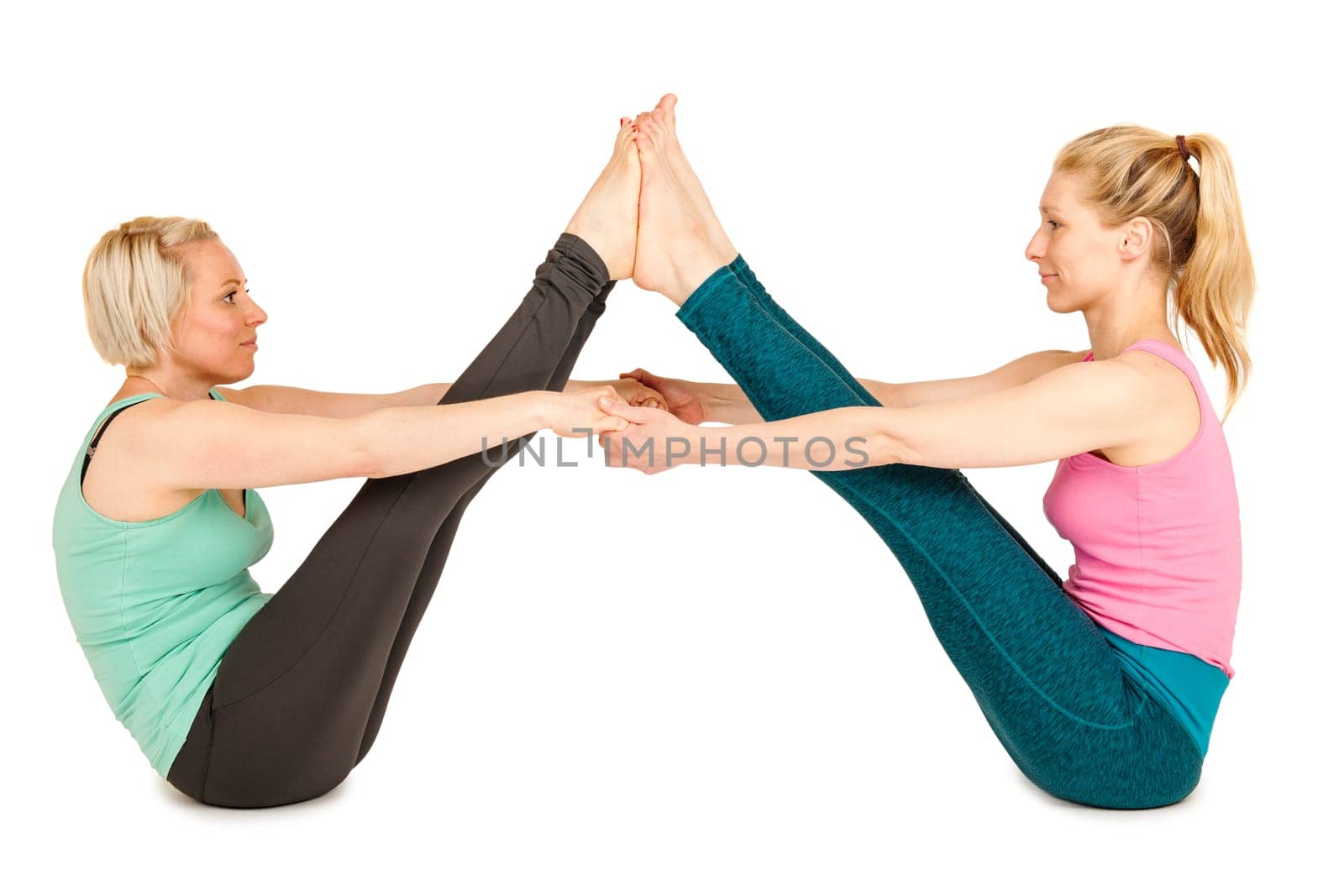Side full-body view of two young blonde women sitting on the floor,touching each other on the soles of their feet and holding hands during a partner yoga exercise against a white background with slight shadows.