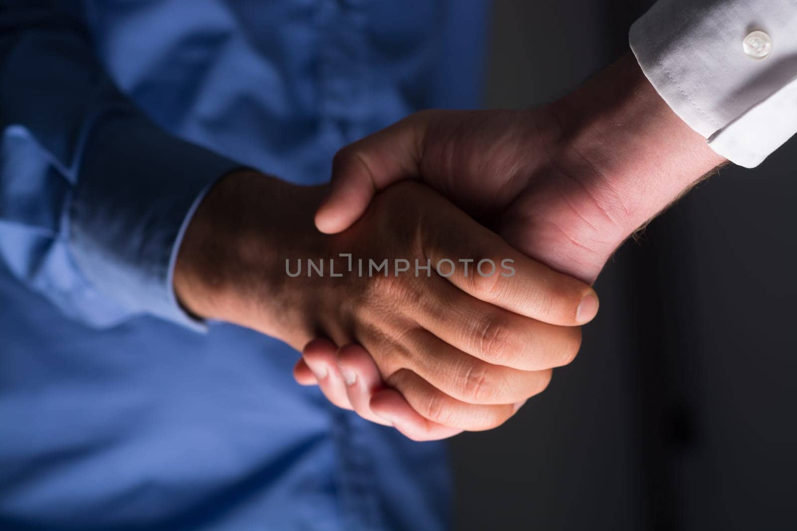 Low angle view of two businessmen hands shaking over dark gray background and light source at bottom side