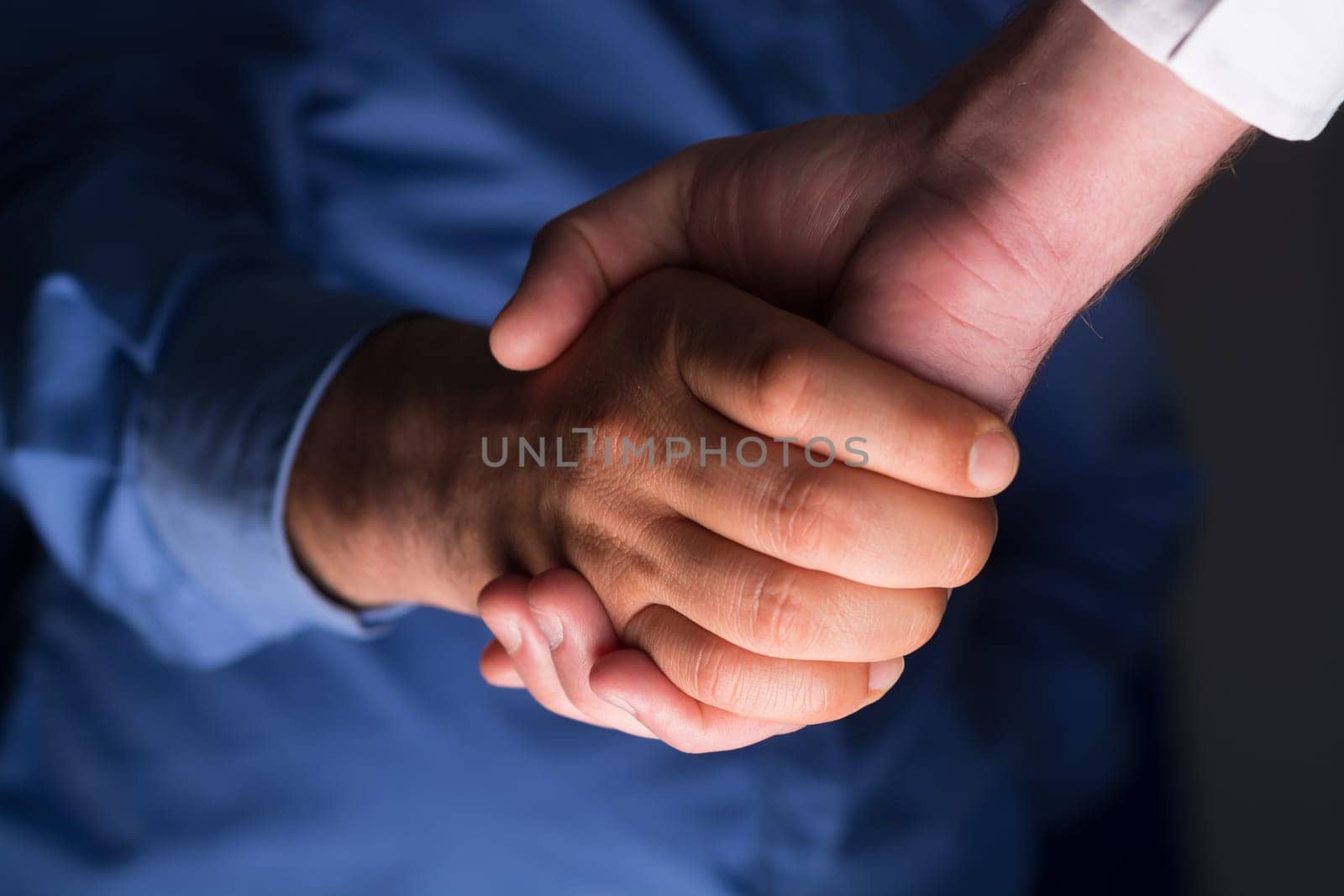 Low angle view of two businessmen hands shaking over dark gray background and light source at bottom side
