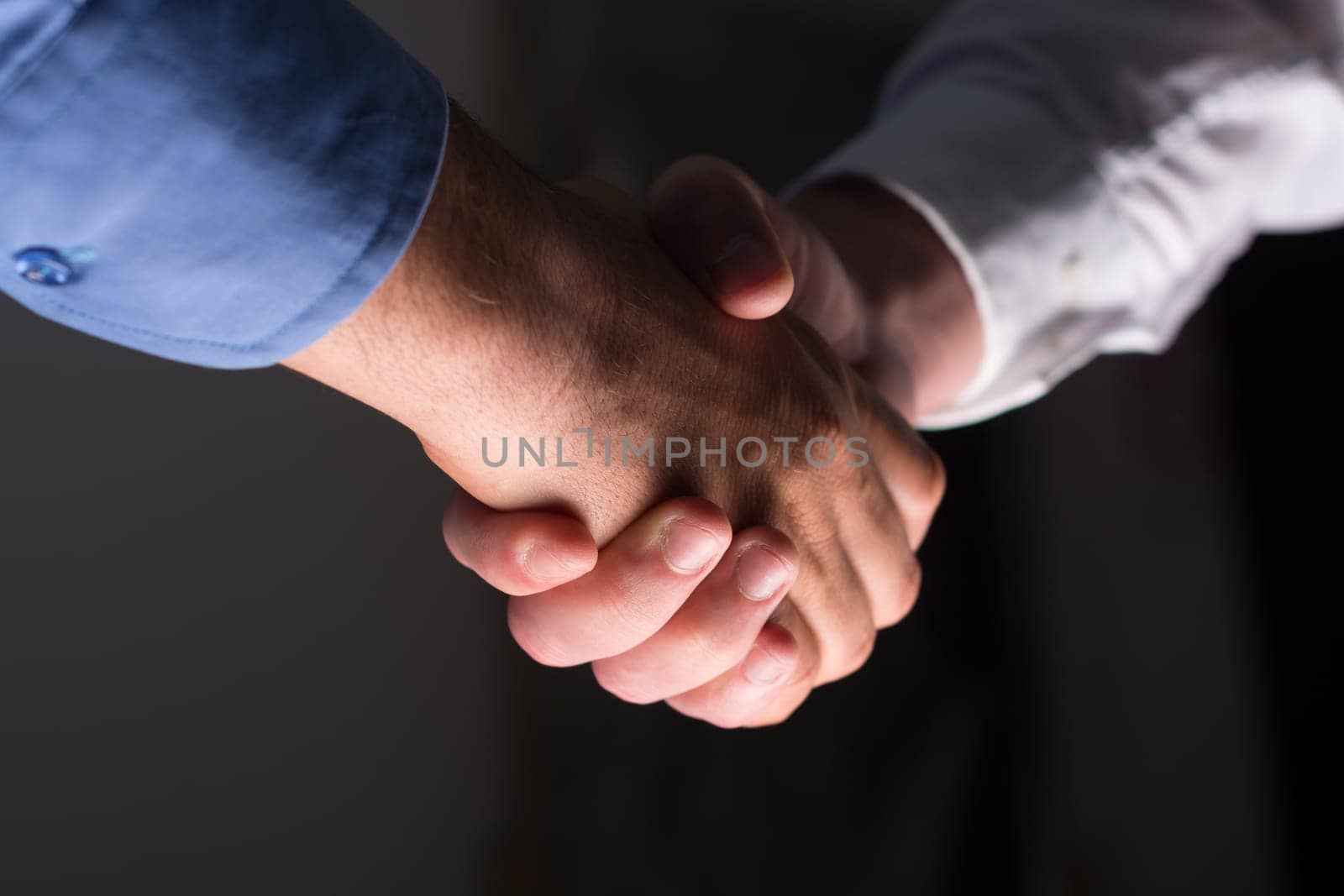 Low angle view of two businessmen hands shaking over dark gray background and light source at bottom side