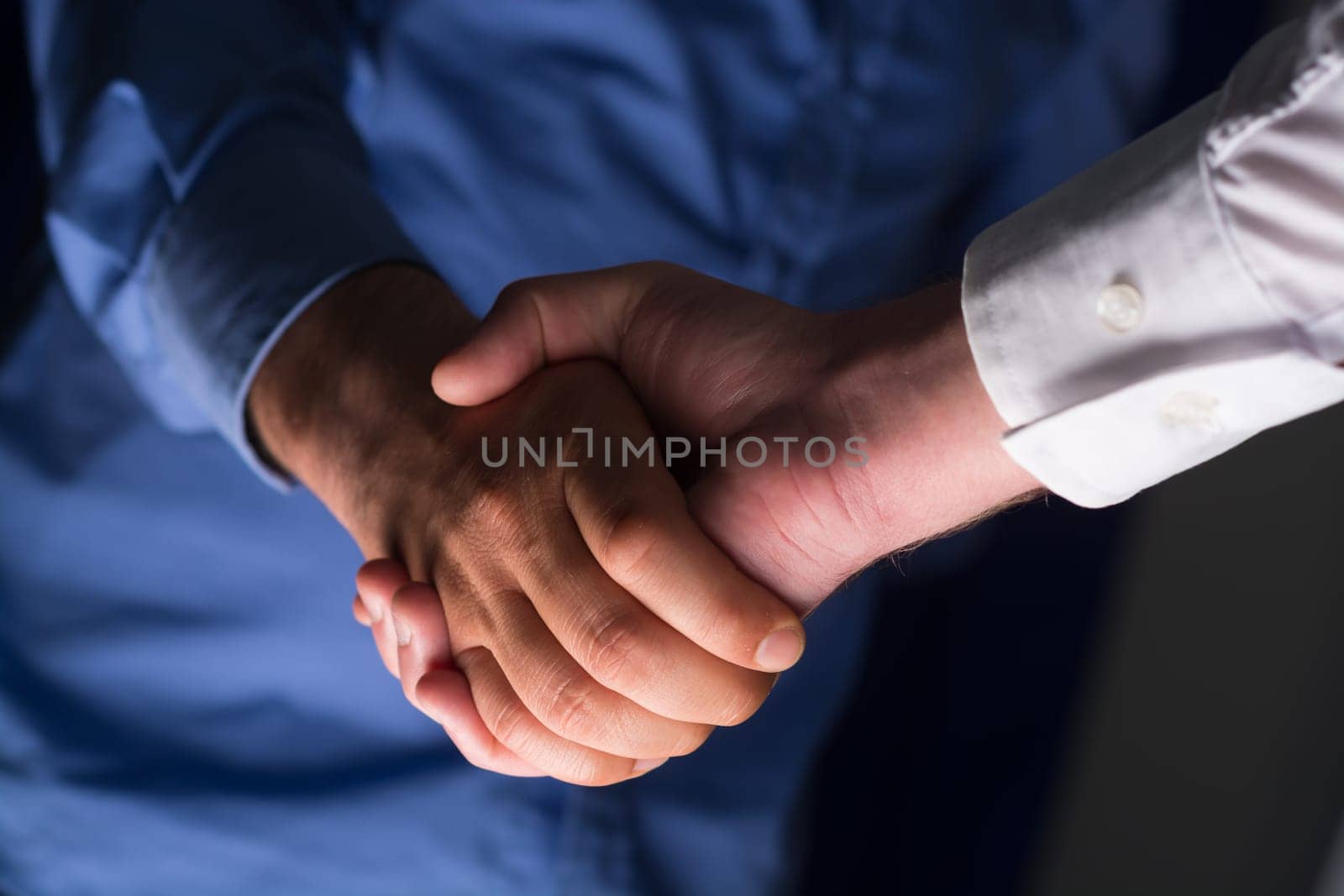 Low angle view of two businessmen hands shaking over dark gray background and light source at bottom side