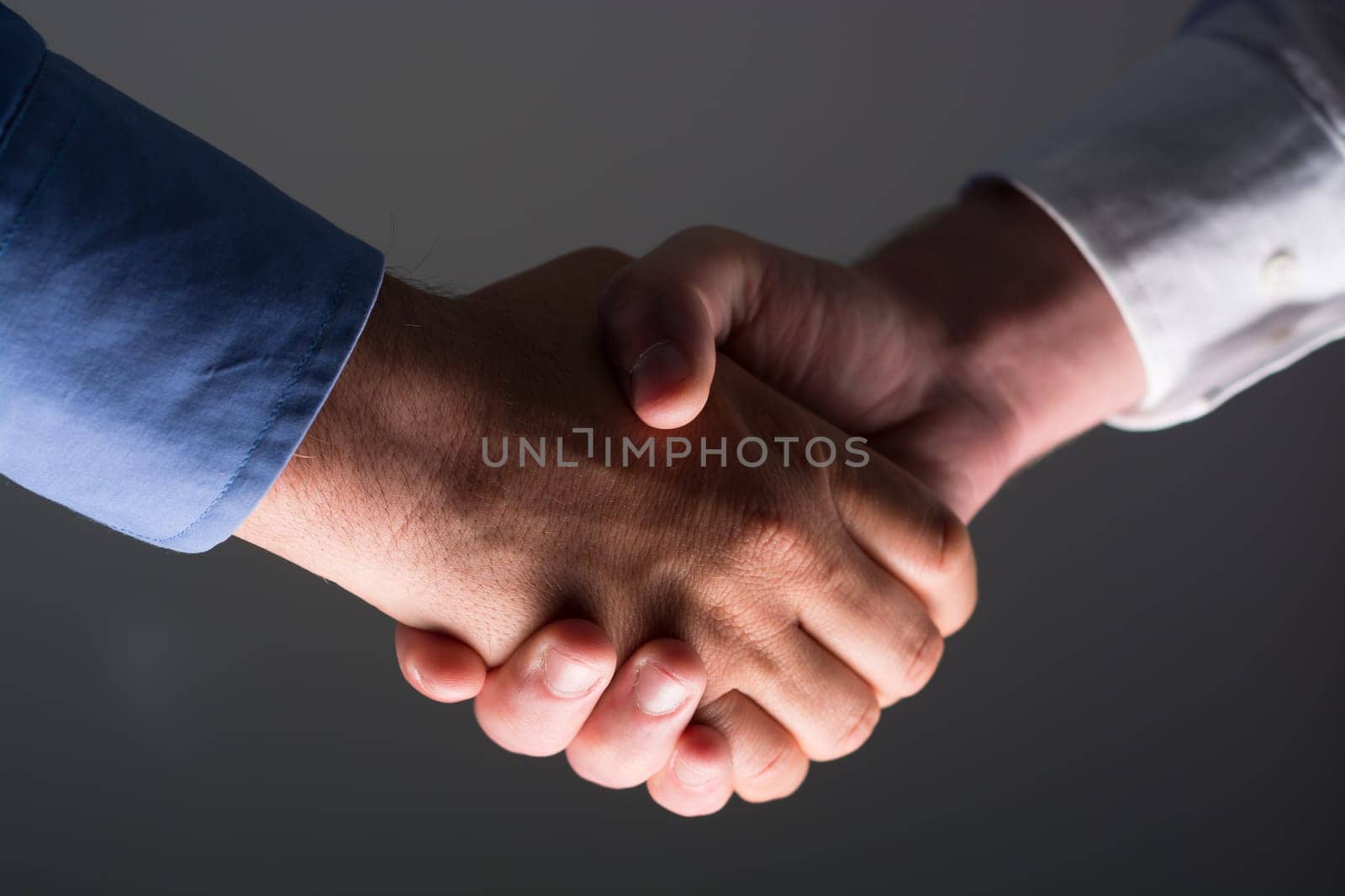 Low angle view of two businessmen hands shaking over dark gray background and light source at bottom side
