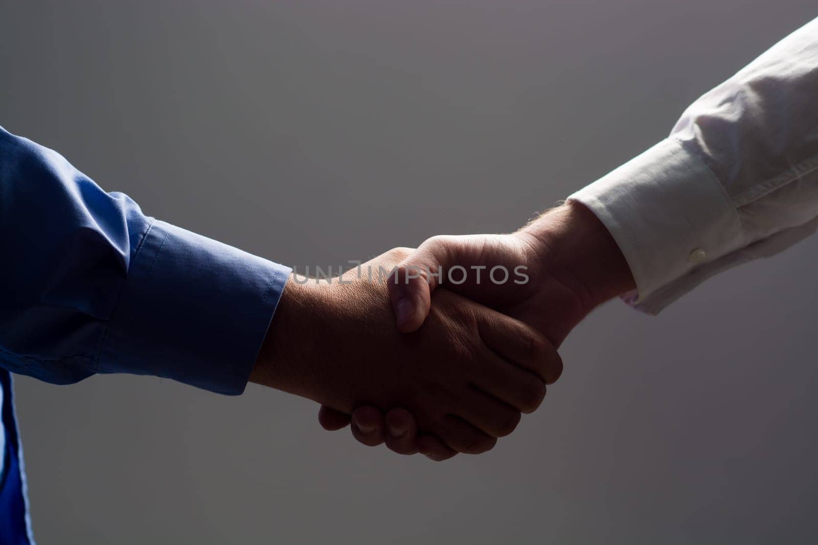 Low angle view of two businessmen hands shaking over dark gray background and light source at bottom side