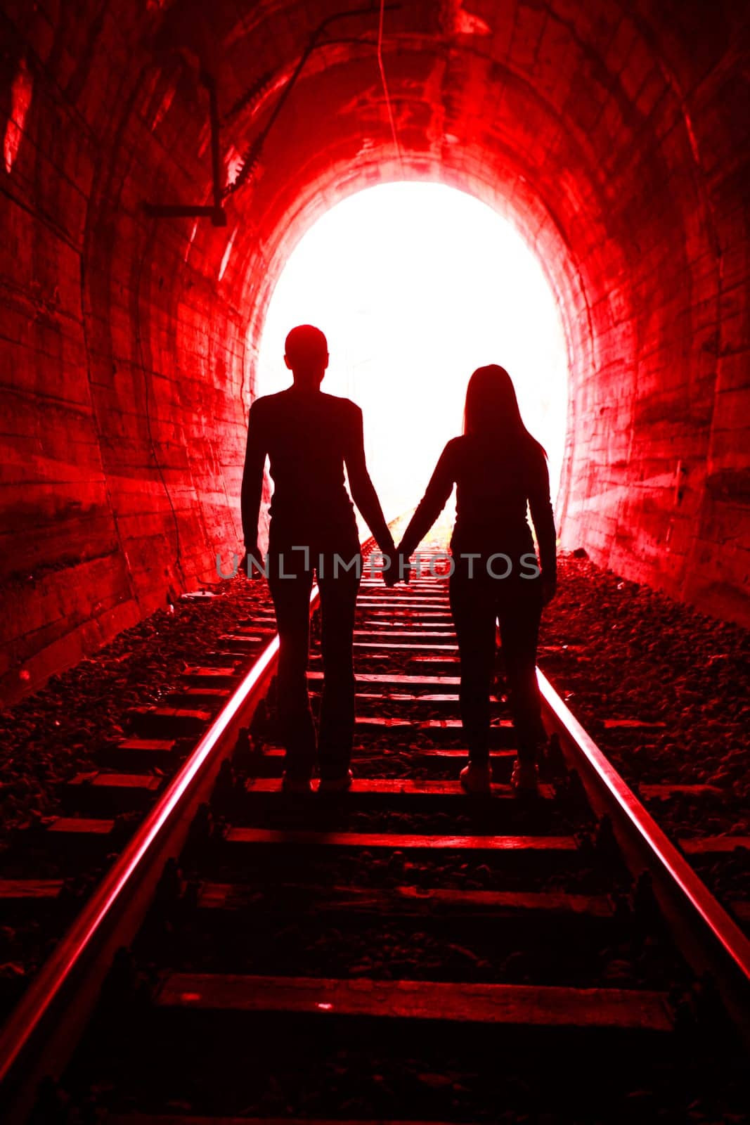 Couple walking hand in hand along the track through a railway tunnel towards the bright light at the other end, they appear as silhouettes against the light