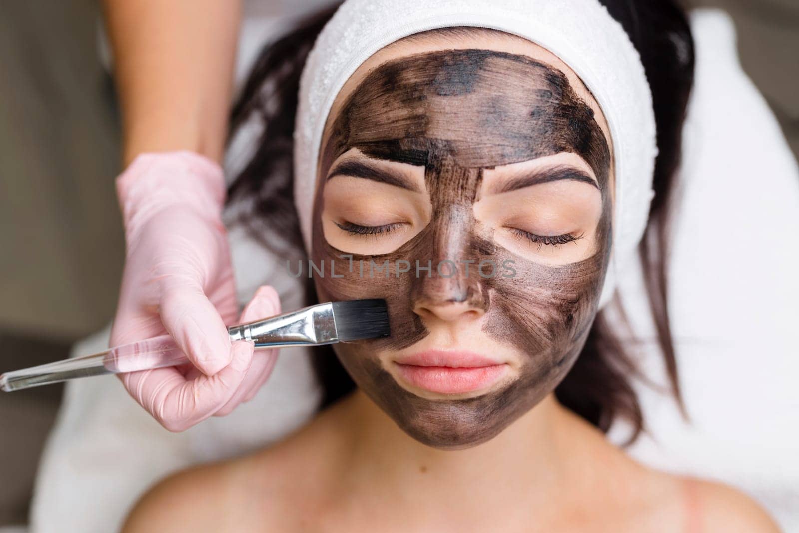cosmetologist applying black mask on pretty woman face wearing black gloves, gorgeous woman in spa having facial procedures.