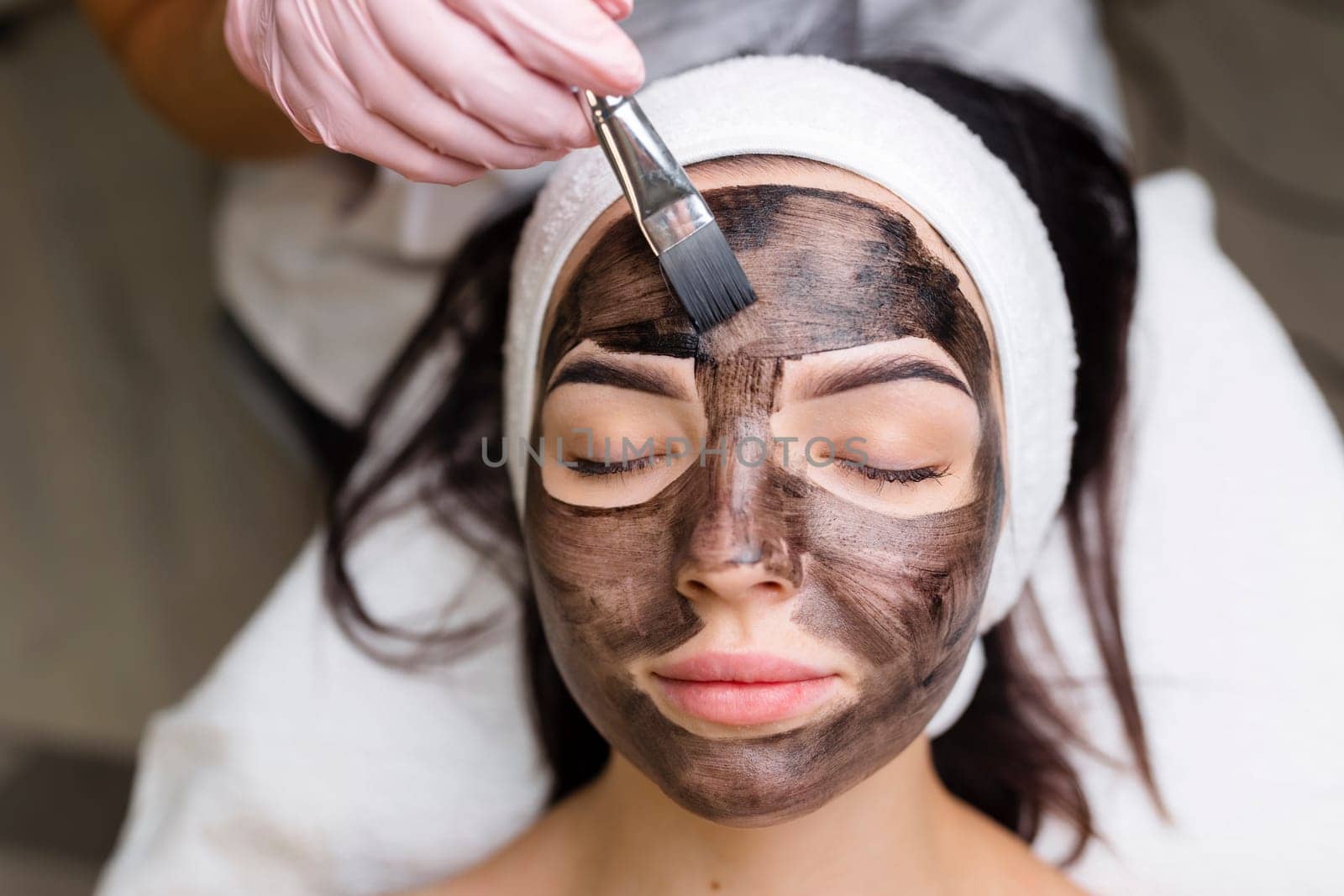 cosmetologist applying black mask on pretty woman face wearing black gloves, gorgeous woman in spa having facial procedures.