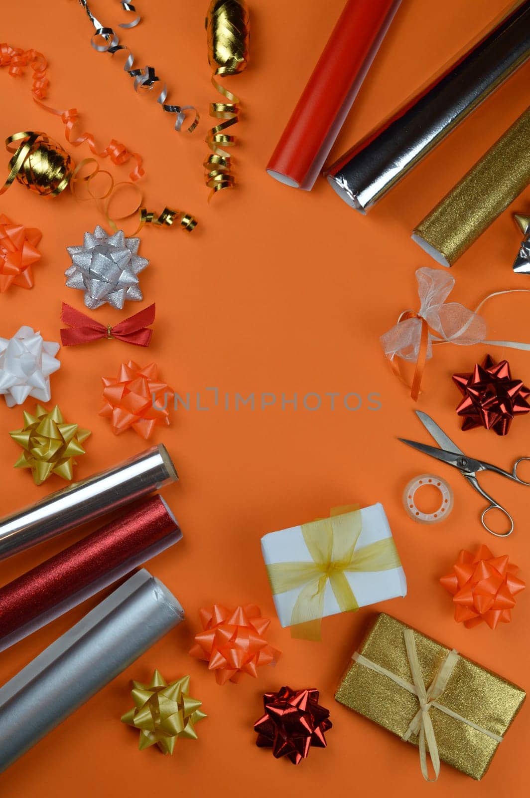 gift wrapping utensils on orange ground in autumn colors red, orange, golden, silver and white with rolls of wrapping paper, gift ribbons, bow topper, scissors and cellotape