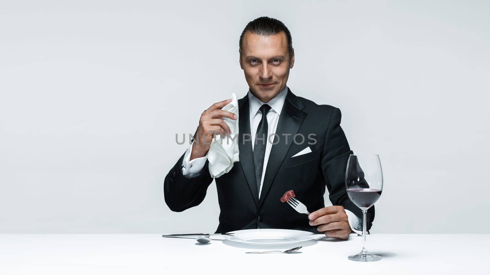 Portrait of a young man in image of maniak with knife, fork and and a piece of raw meat on a plate. Man in suit. The image in the style of Halloween