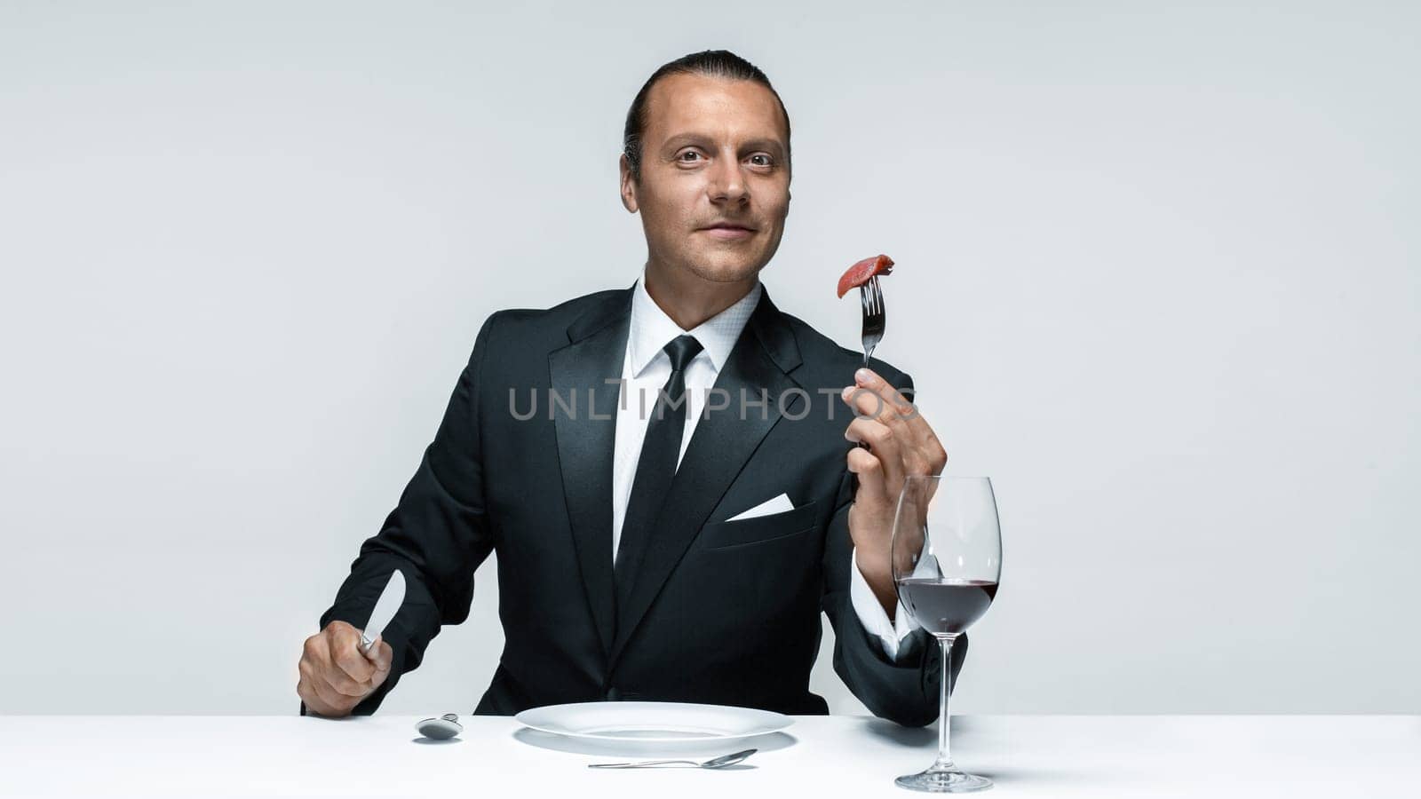Portrait of a young man in image of maniak with knife, fork and and a piece of raw meat on a plate. Man in suit. The image in the style of Halloween