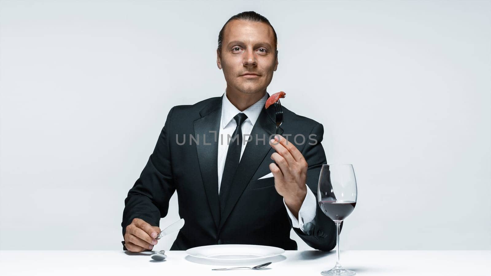 Portrait of a young man in image of maniak with knife, fork and and a piece of raw meat on a plate. Man in suit. The image in the style of Halloween