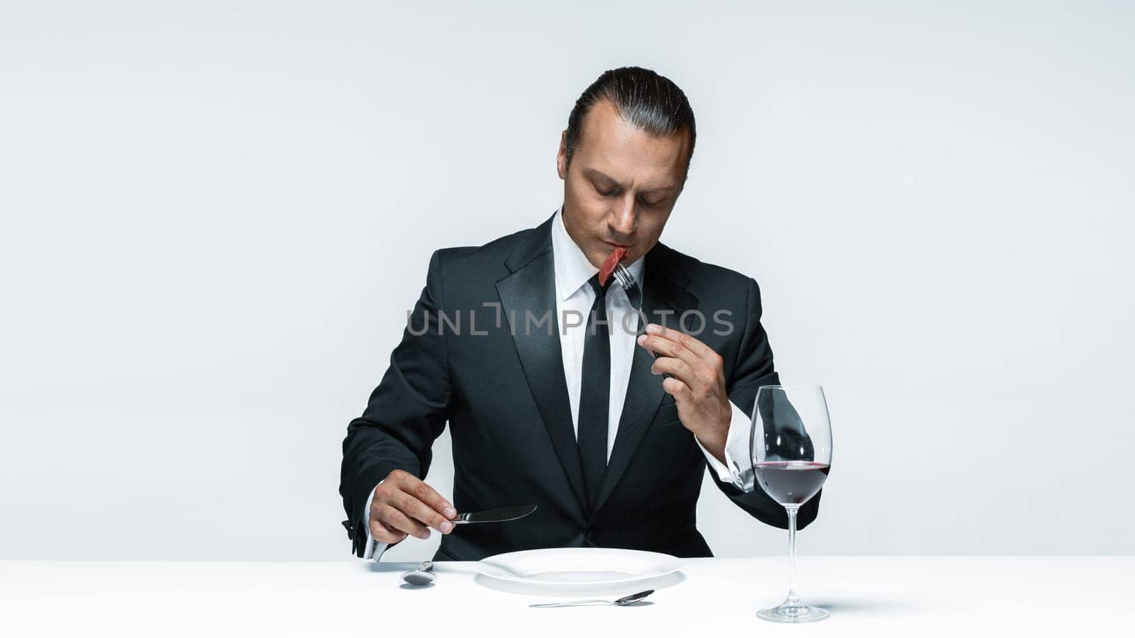 Portrait of a young man in image of maniak with knife, fork and and a piece of raw meat on a plate. Man in suit. The image in the style of Halloween