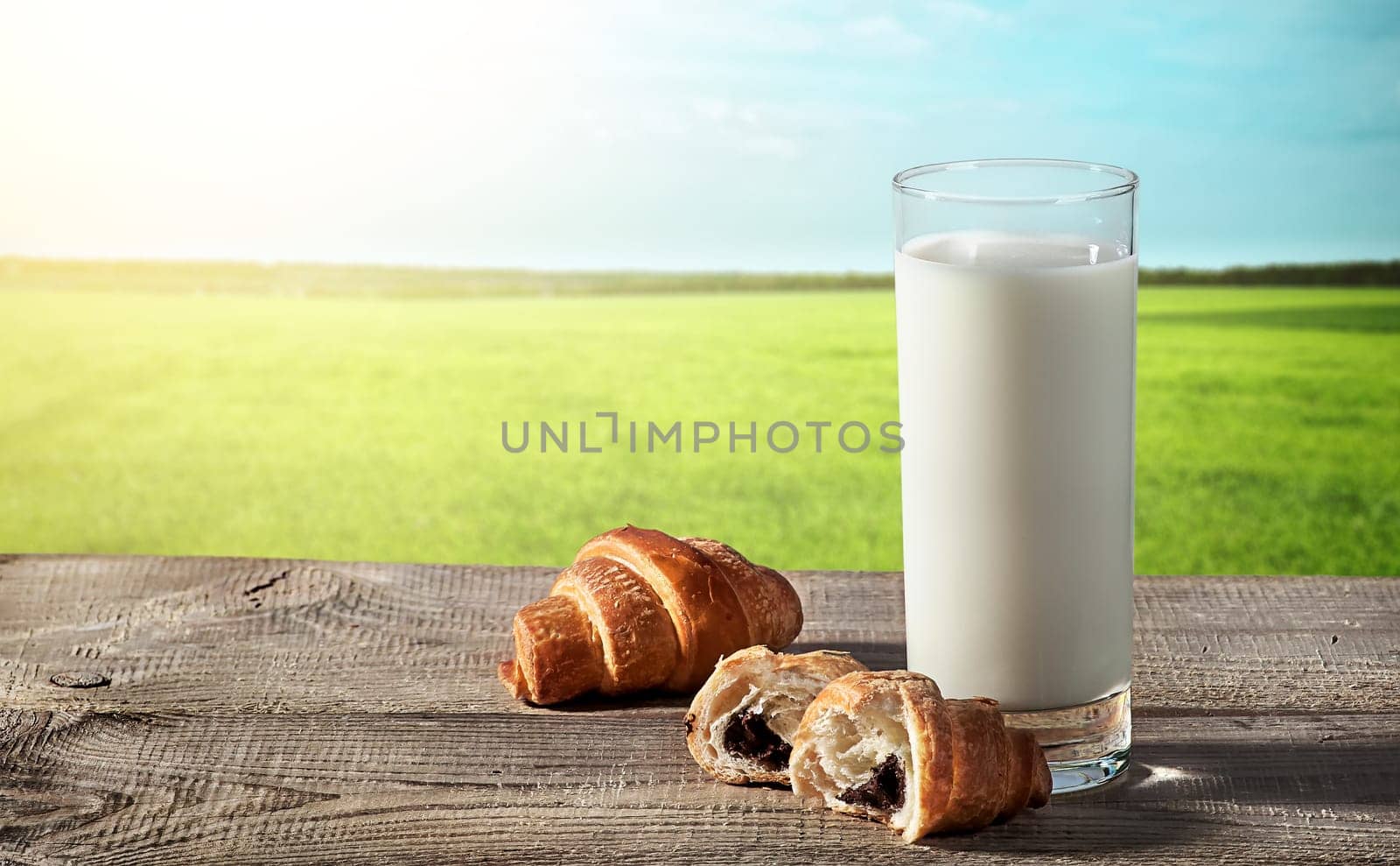 Glass of milk on a rustic table, beside croissants, blurred background with grass and sky