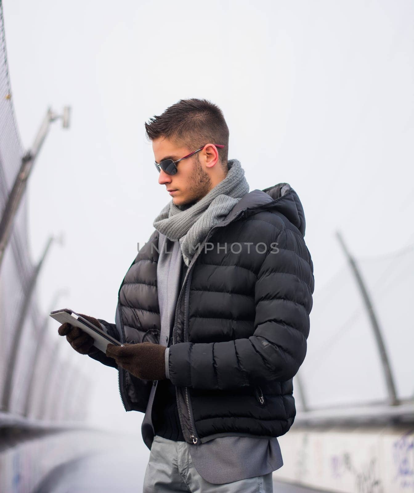 Handsome unshaven trendy man in winter fashion wearing a thick jacket, scarf and sunglasses standing looking down at a tablet computer that he is holding, side view