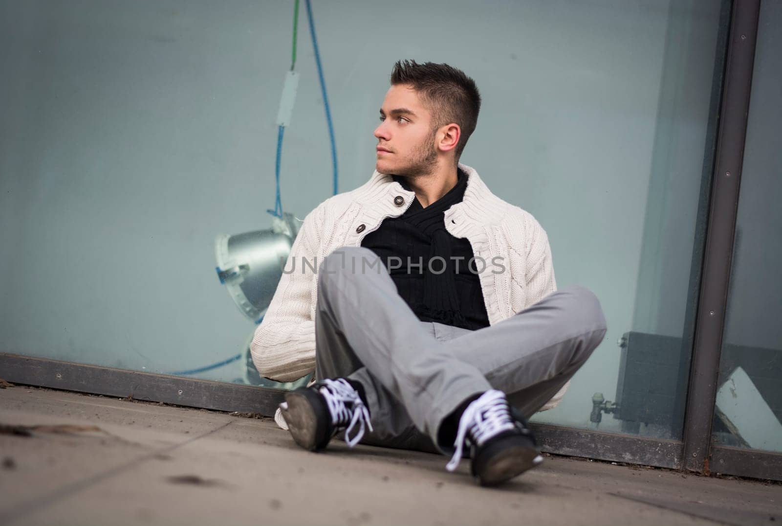Attractive young man sitting against abandoned shop window, looking away to a side