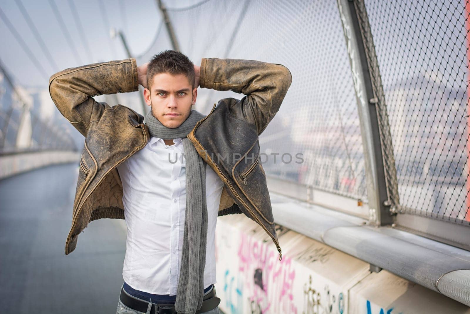 Handsome trendy young man standing on a sidewalk wearing a fashionable jacket and scarf with arms raised behind head looking at the camera