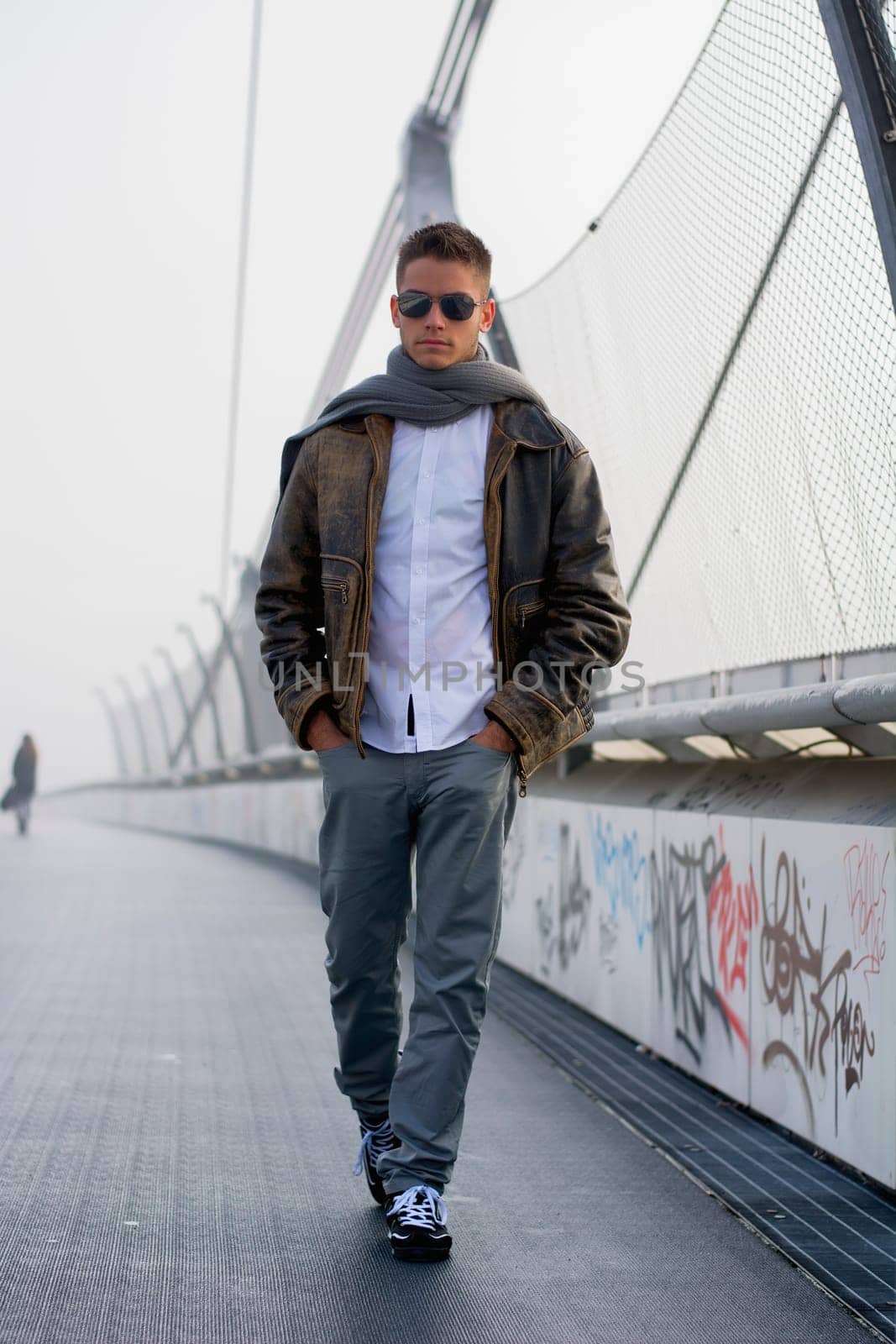 Handsome trendy young man standing on a sidewalk wearing a fashionable leather jacket and scarf in a relaxed confident pose looking at the camera