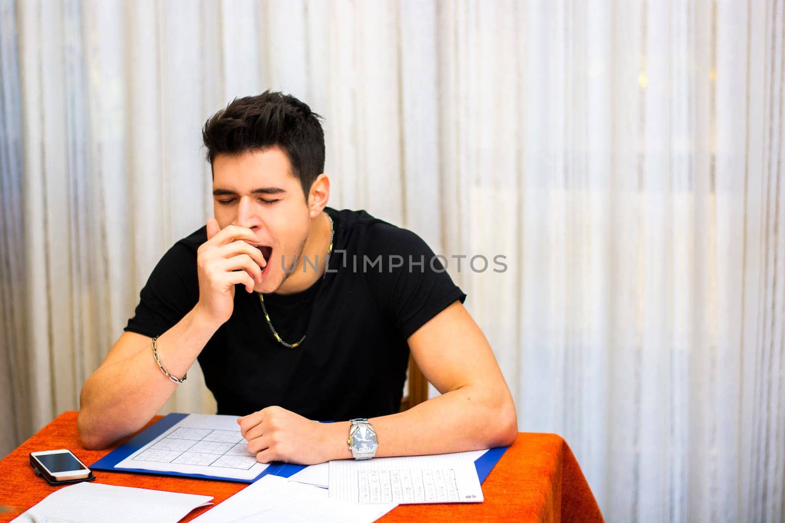Tired, exhausted young man yawning while working or studying at home sitting at table with paper sheets in front of him