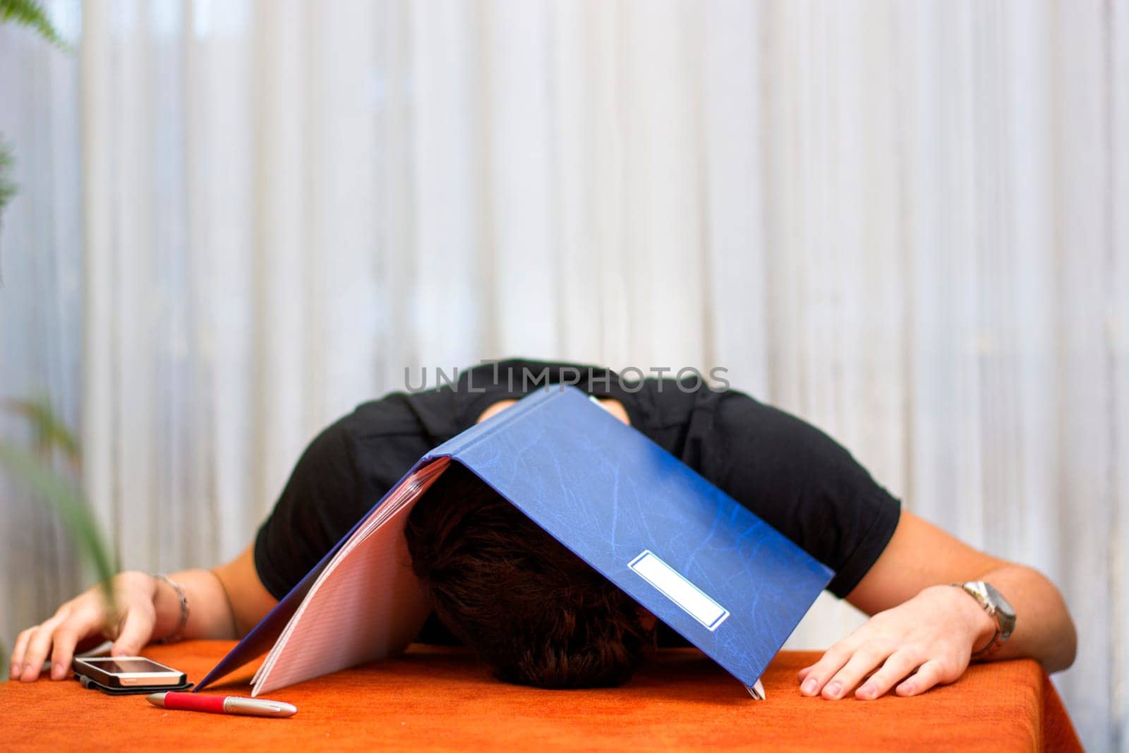 Tired, exhausted or despondent young man sitting at a table with a folder of paperwork over his head