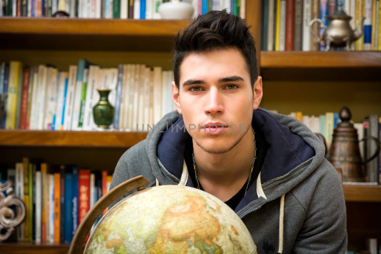 Handsome young man holding a globe indoors at home looking at the camera in front of a bookcase filled with books in a conceptual image