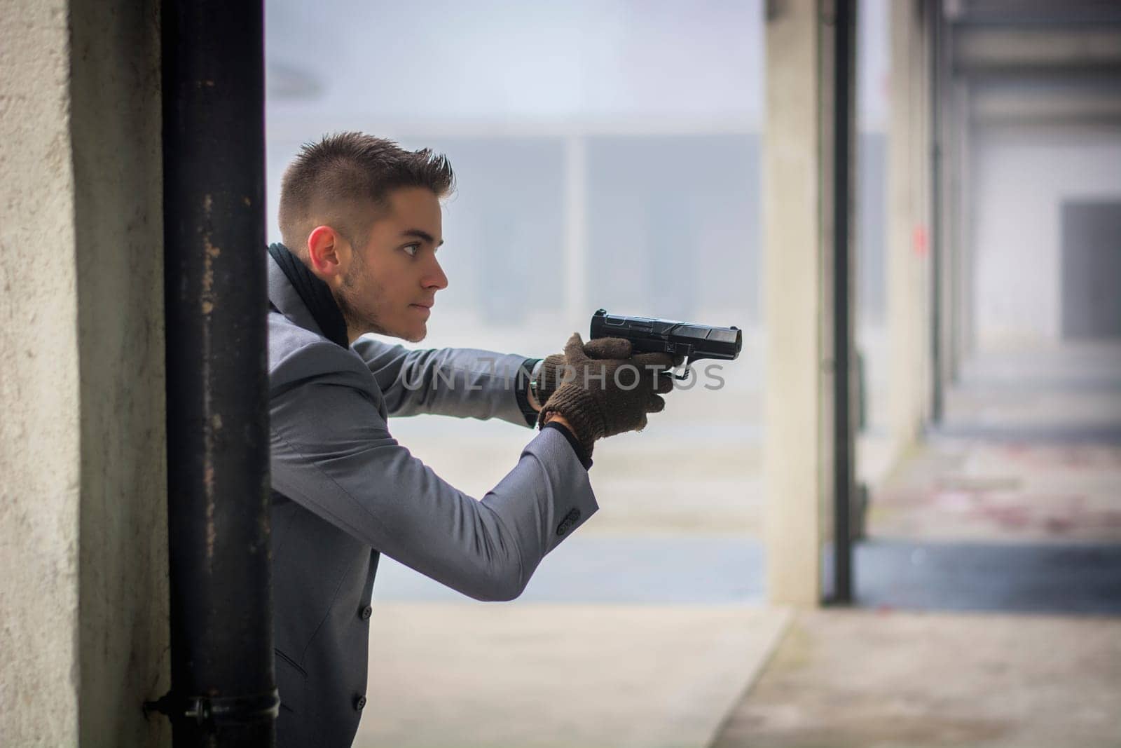 Well dressed handsome young detective or policeman or mobster standing in an urban environment holding a gun with a determined expression, coming from behind a column