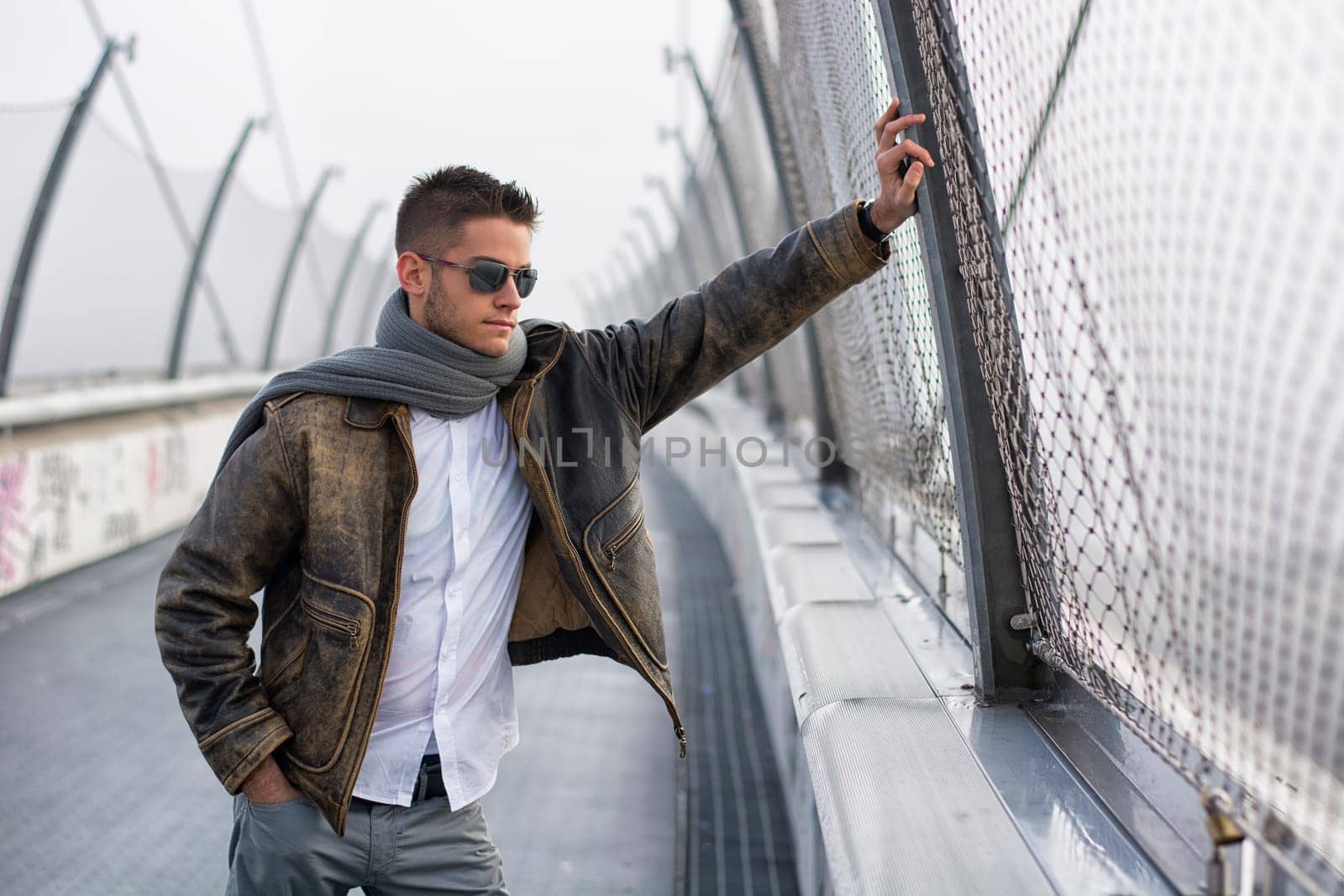 Handsome trendy young man standing on a sidewalk wearing a fashionable jacket and scarf in a relaxed confident pose looking away down the bridge