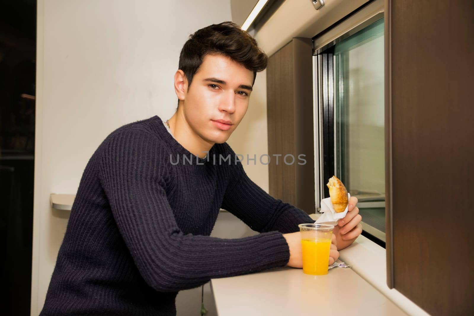 Young man eating refreshments on a train looking at camera while holding a pastry, with orange juice on the ledge below the window, while commuting or traveling