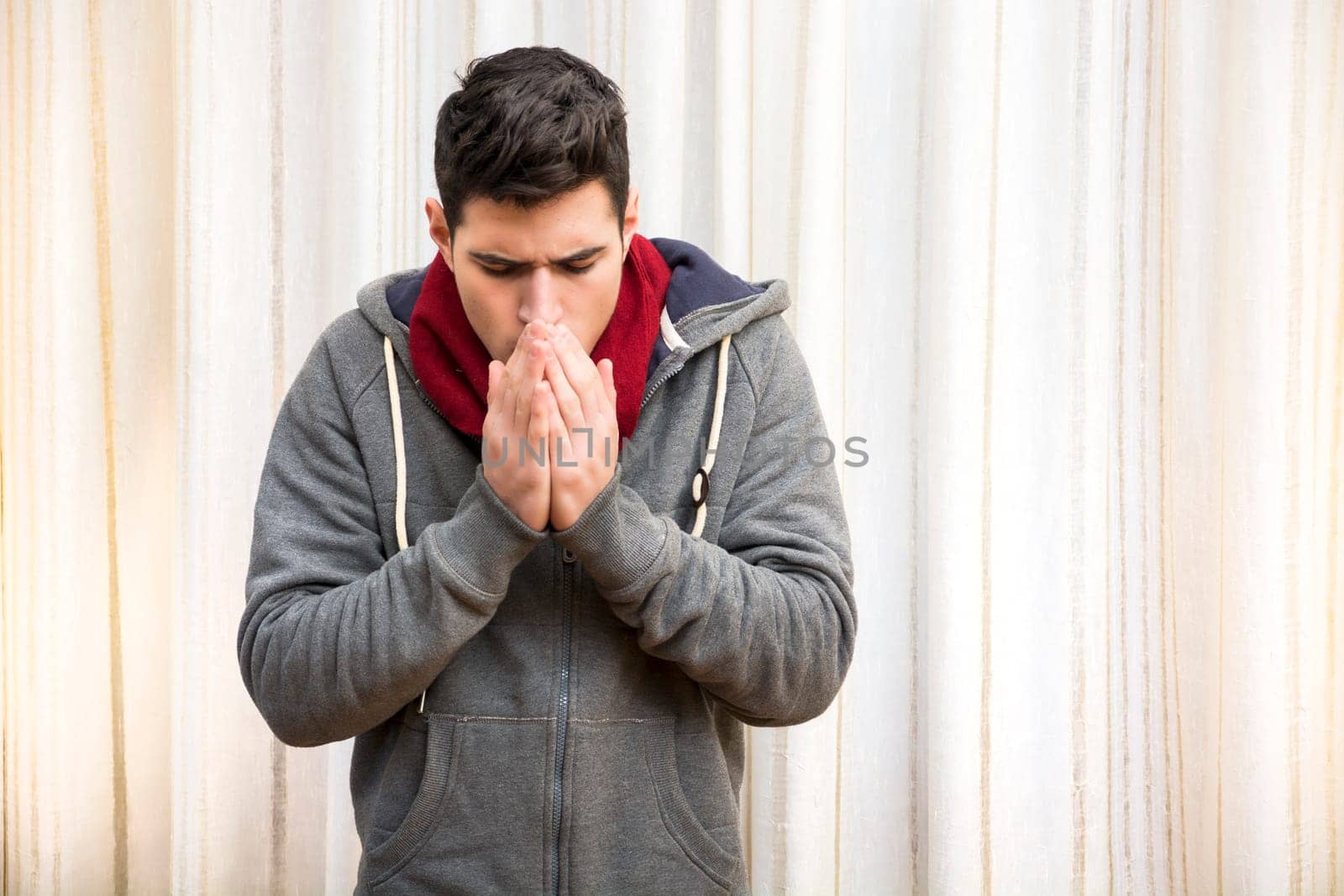 Young man feeling very cold, warming hands with his breath, wearing heavy sweater and scarf