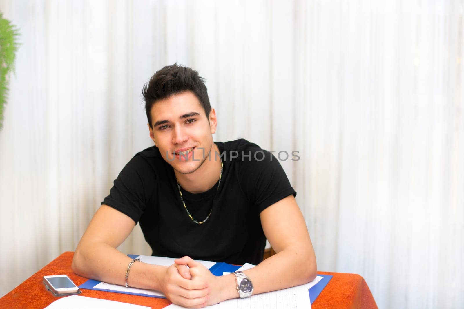 Attractive smiling young man working or studying at home sitting at table with paper sheets in front of him