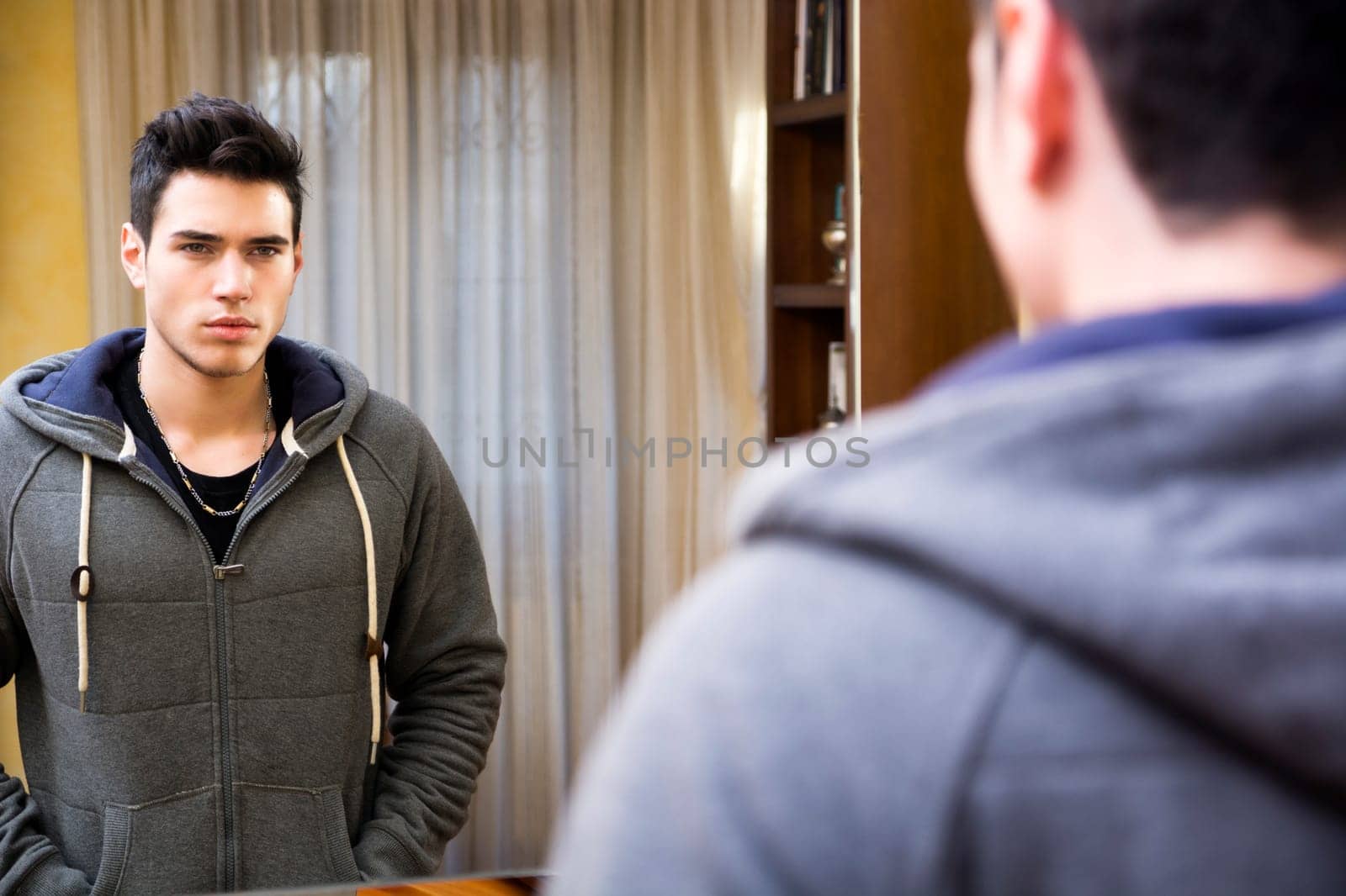 Handsome young man looking at his reflection in the mirror in living room at home