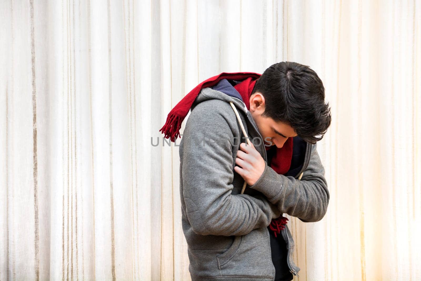 Young man feeling very cold, curling up inside his heavy sweater, wearing a red scarf