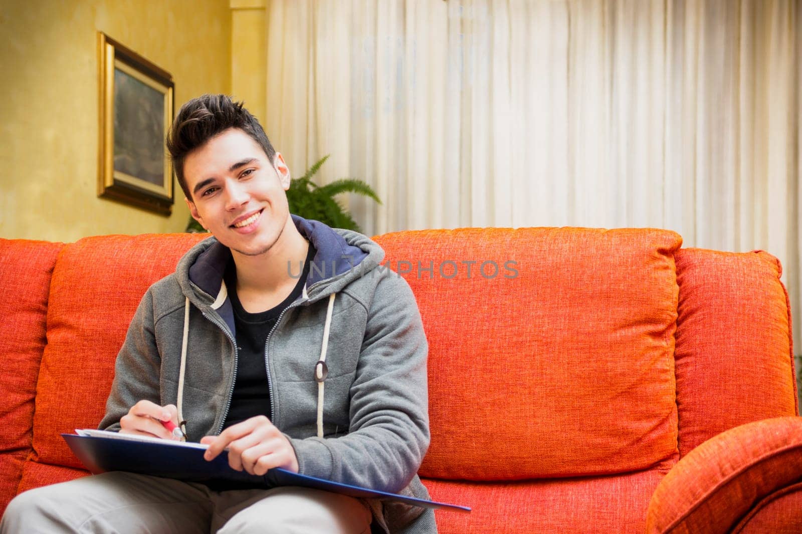 Smiling young man at home writing on notebook, sitting on couch, looking at camera