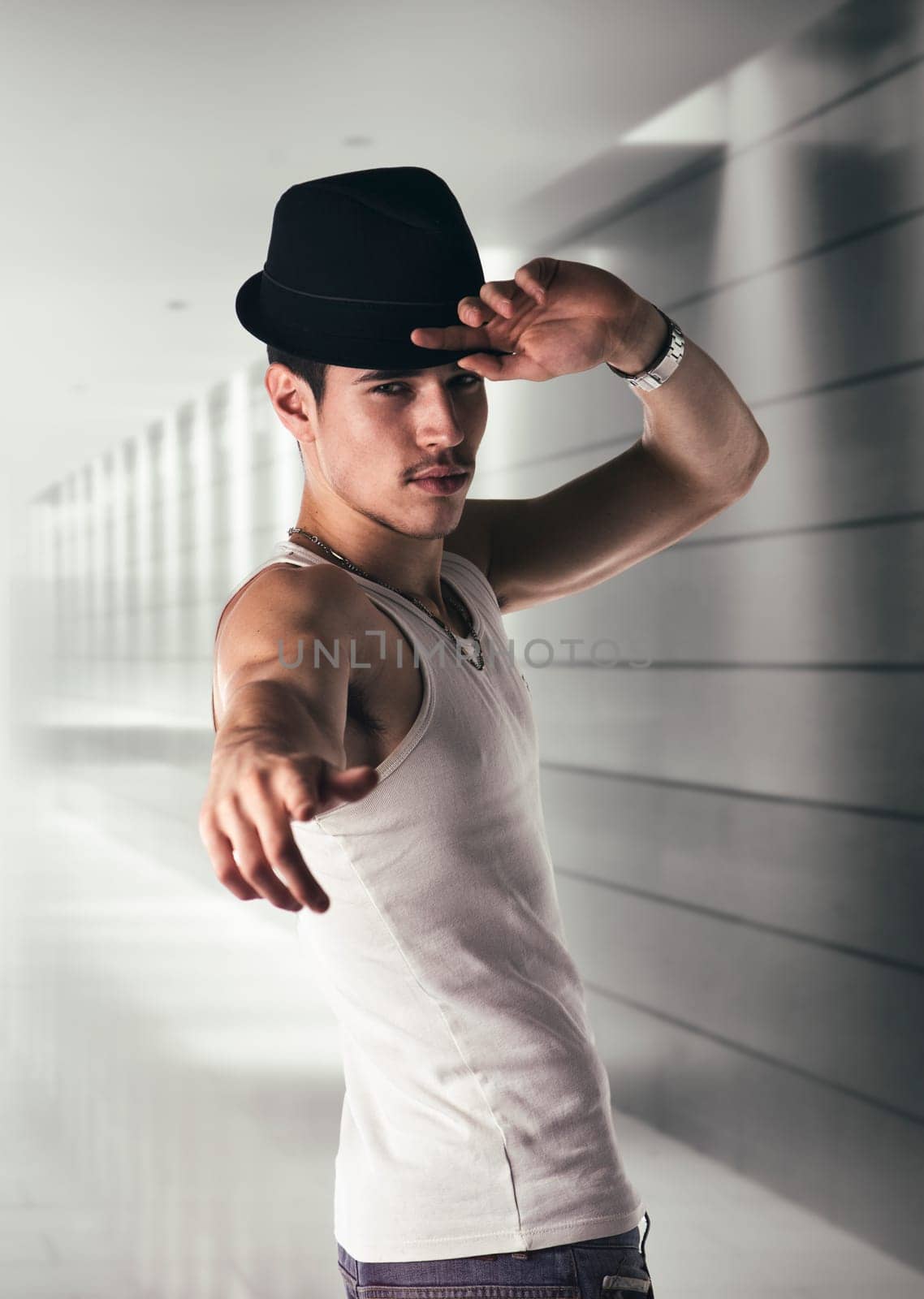 Attractive young man with black fedora hat and white tank-top in a hallway, pointing finger to the camera