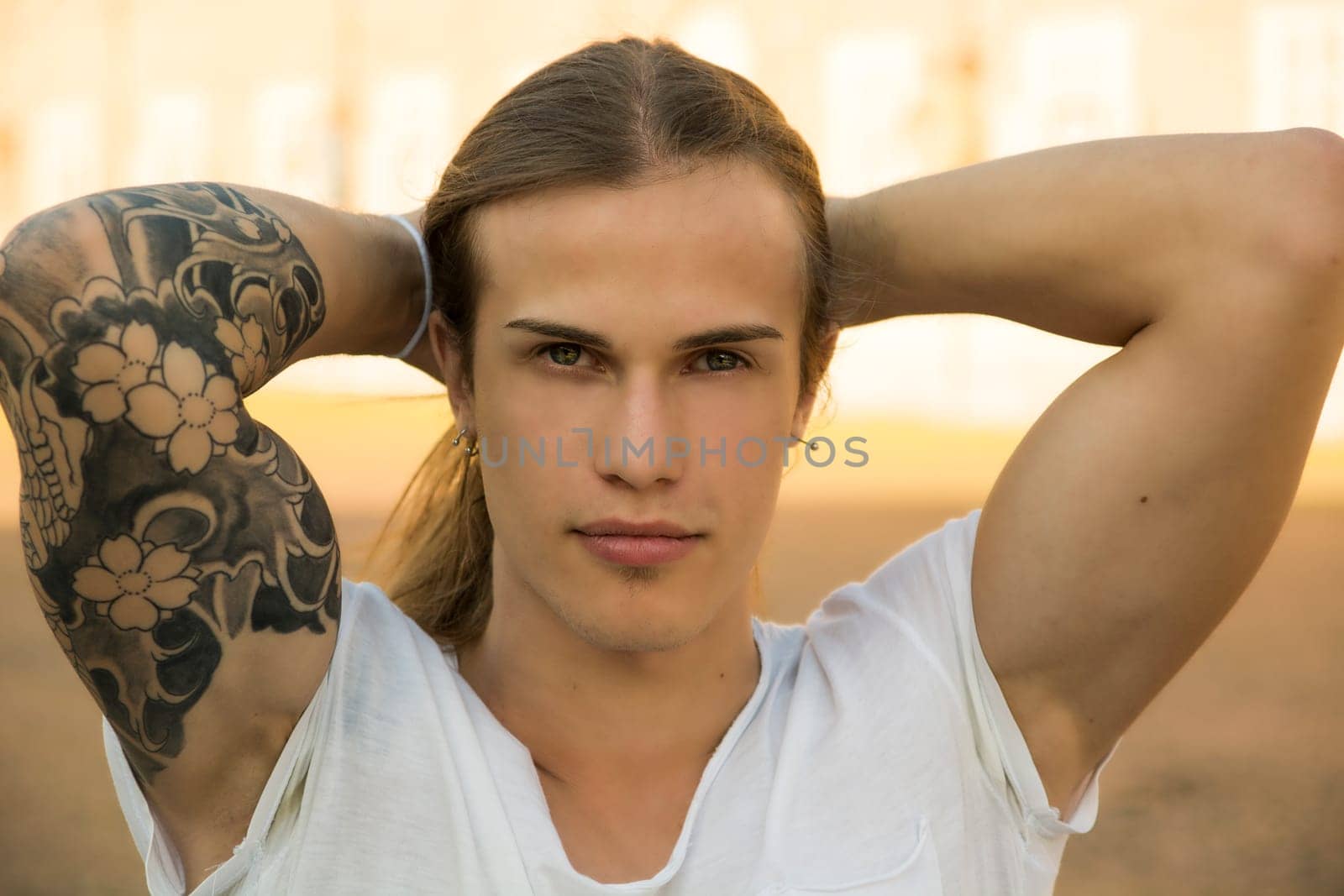 Handsome young man outdoors, with hands behind head, looking intensely at camera with confident expression