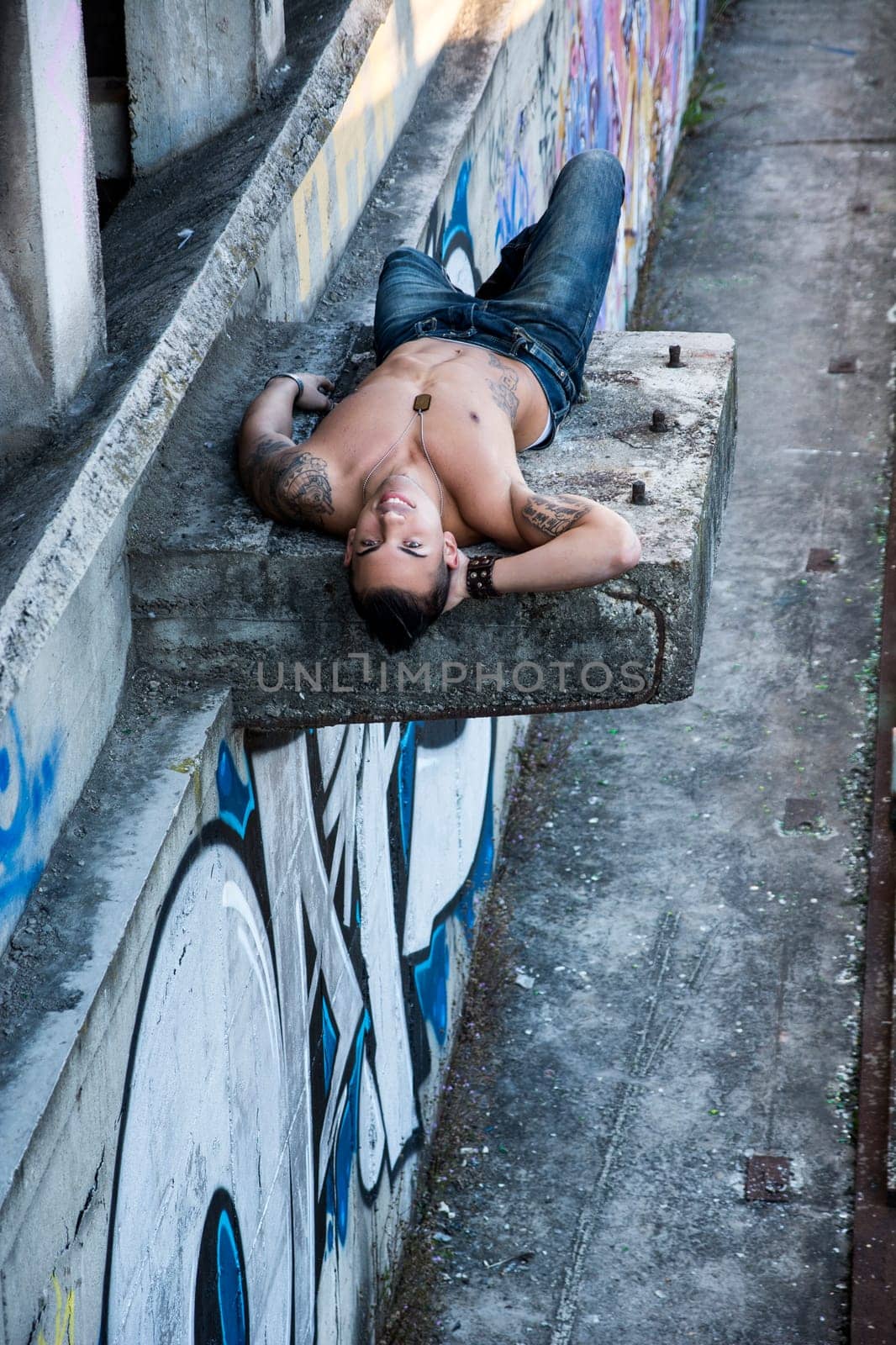 Handsome shirtless young man laying down on concrete structure above the ground, smiling at camera