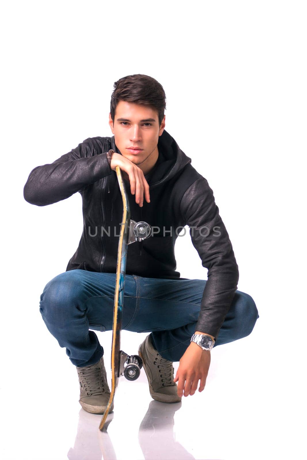 Handsome young man resting head on skateboard sitting on his heels, isolated on white