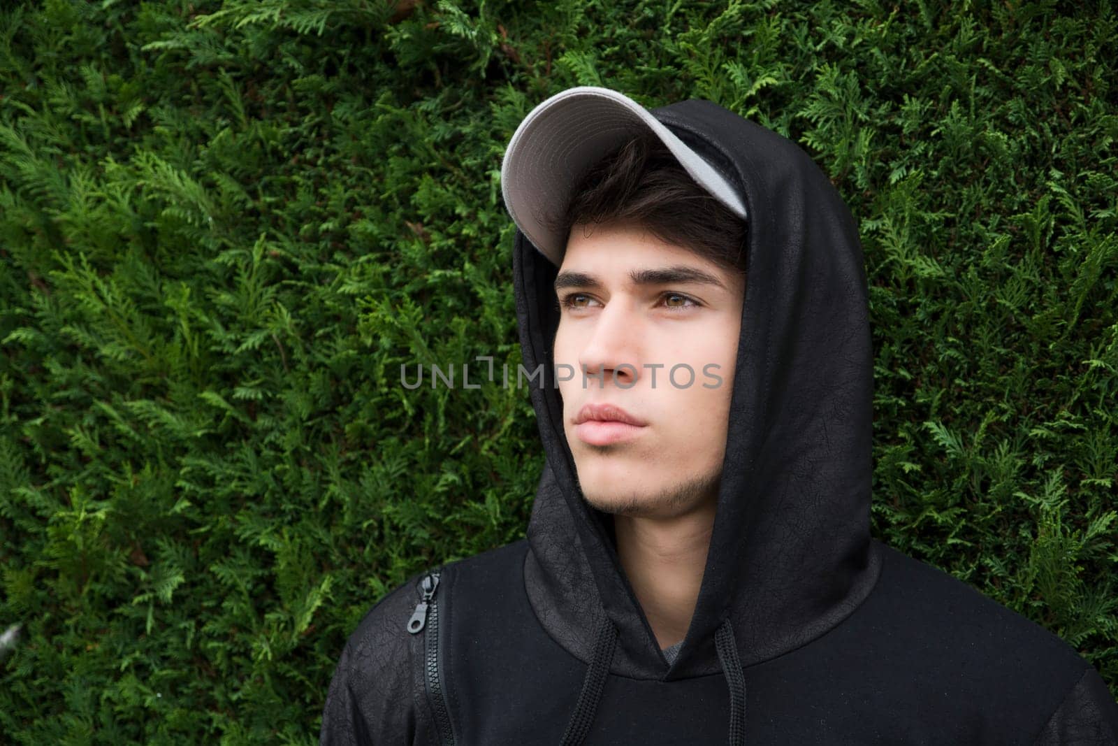 Attractive young man with hoodie and baseball cap against green bush "wall"", sad or thinking"