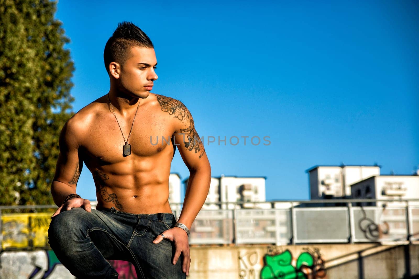 Handsome fit athletic shirtless young man in city setting kneeling and looking to a side, large copyspace