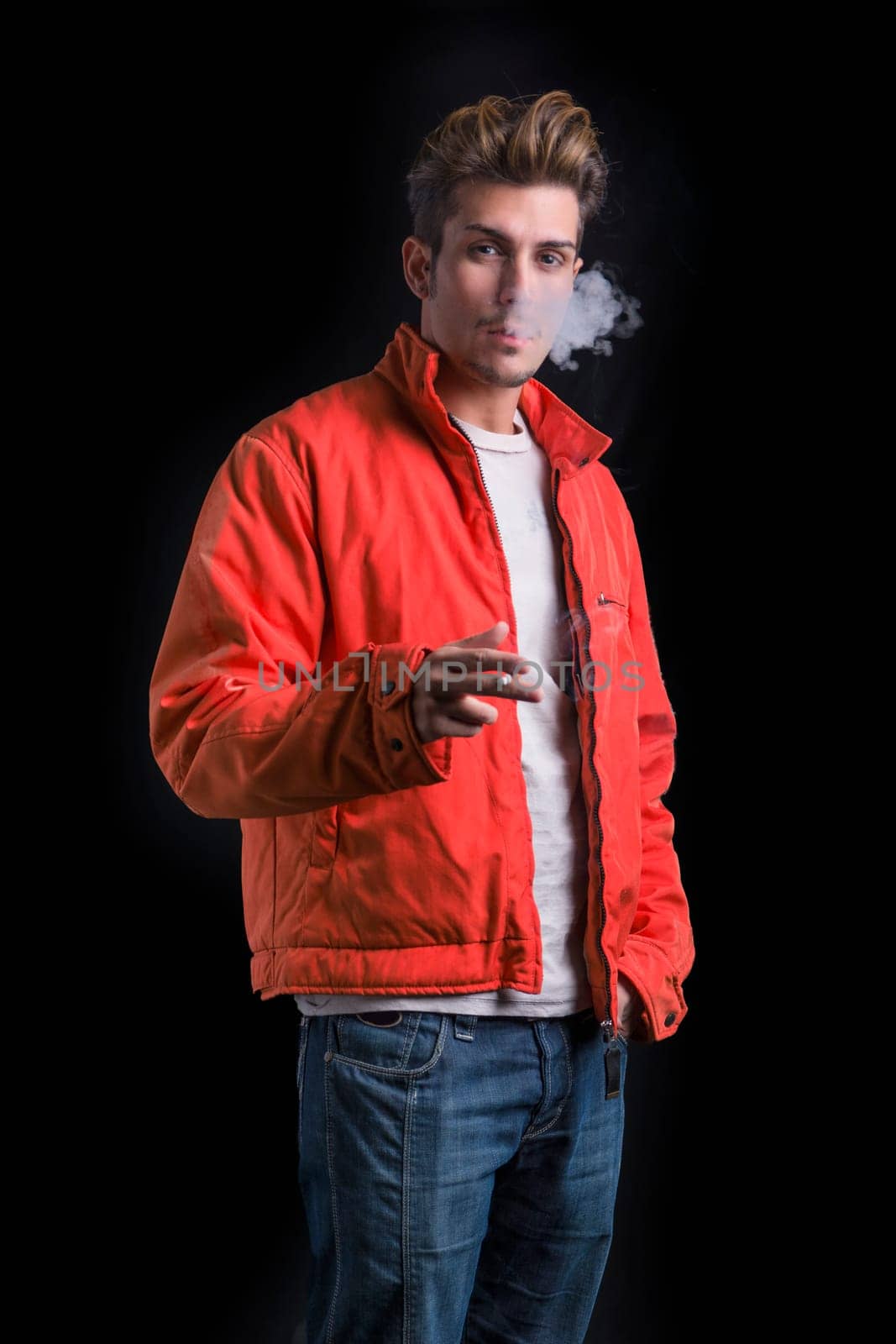 Handsome young man smoking cigarette, dressed as James Dean with red jacket on dark background