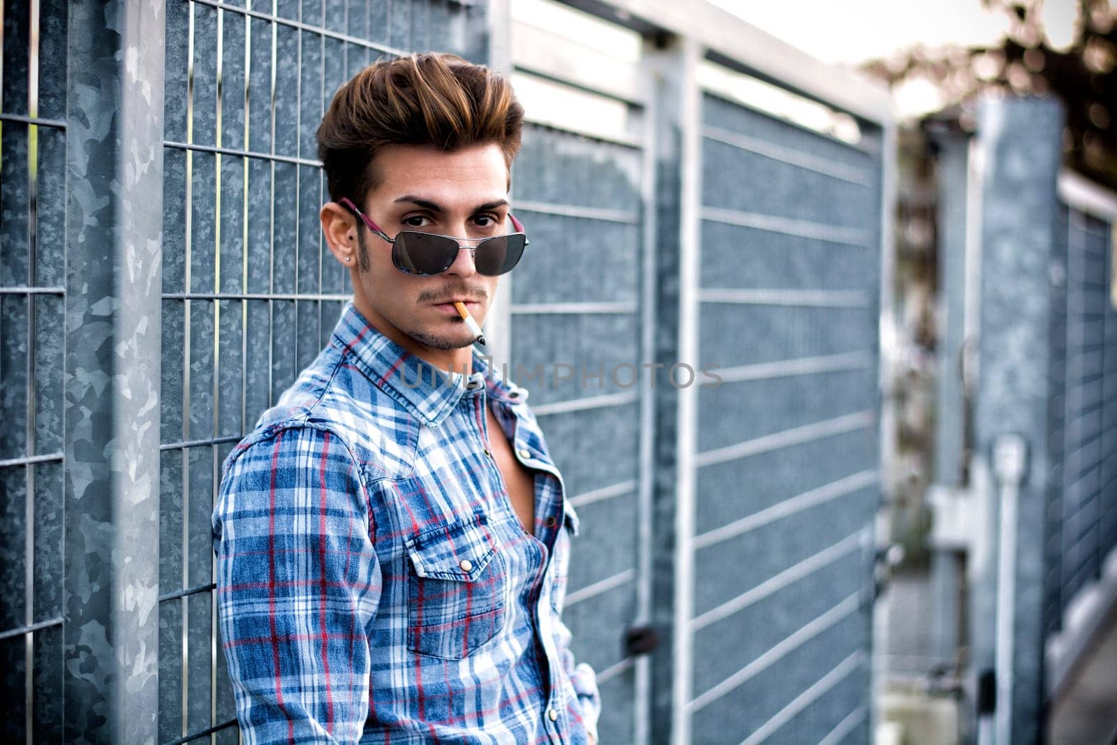 Trendy cool young man standing outside smoking, leaning against metal gate, wearing shirt and jeans