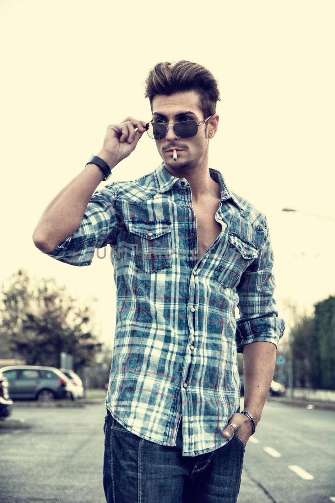 Trendy cool young man standing outside smoking, lowering sunglasses, wearing shirt and jeans