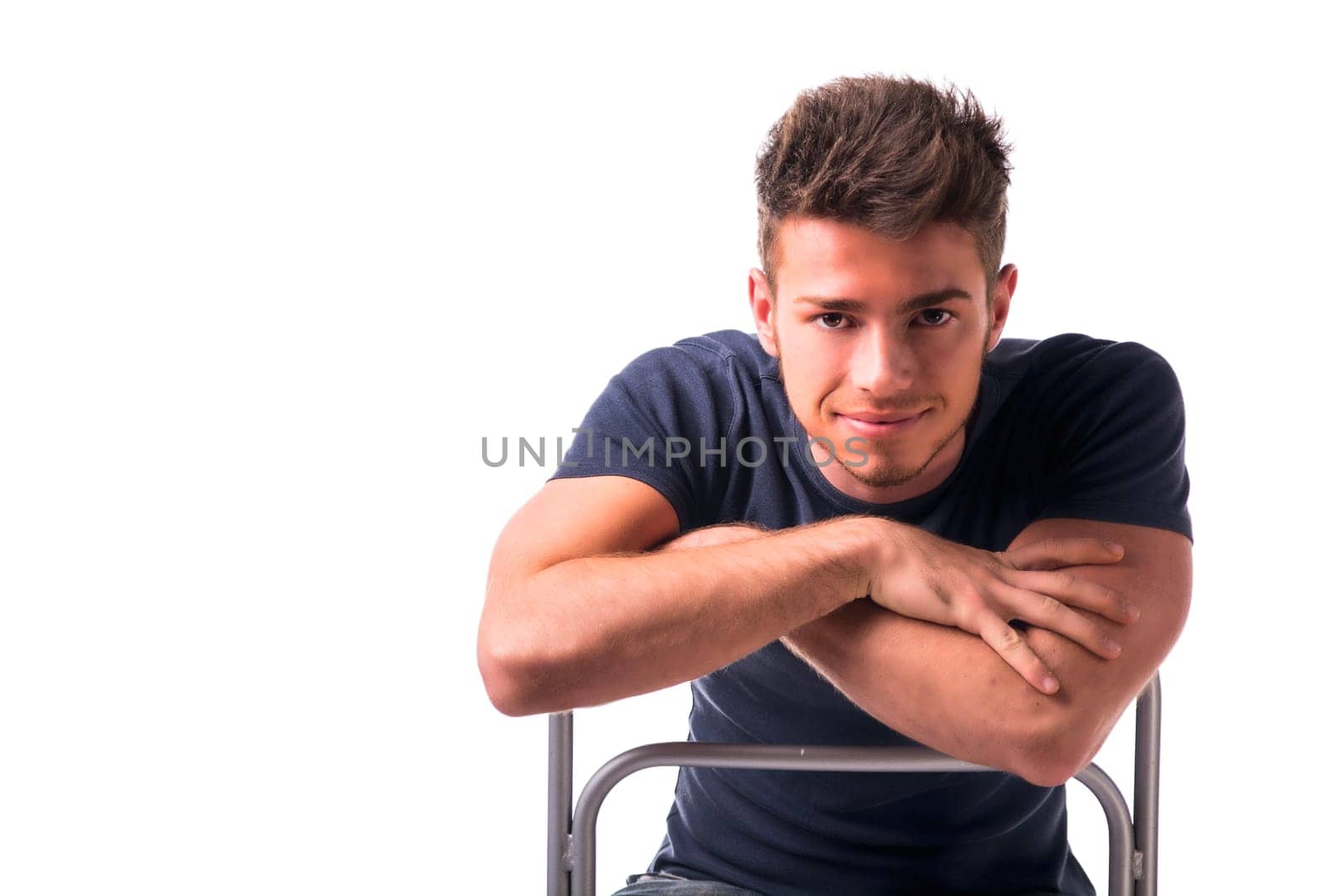Attractive young man sitting, leaning on back of a chair looking at camera, isolated on white