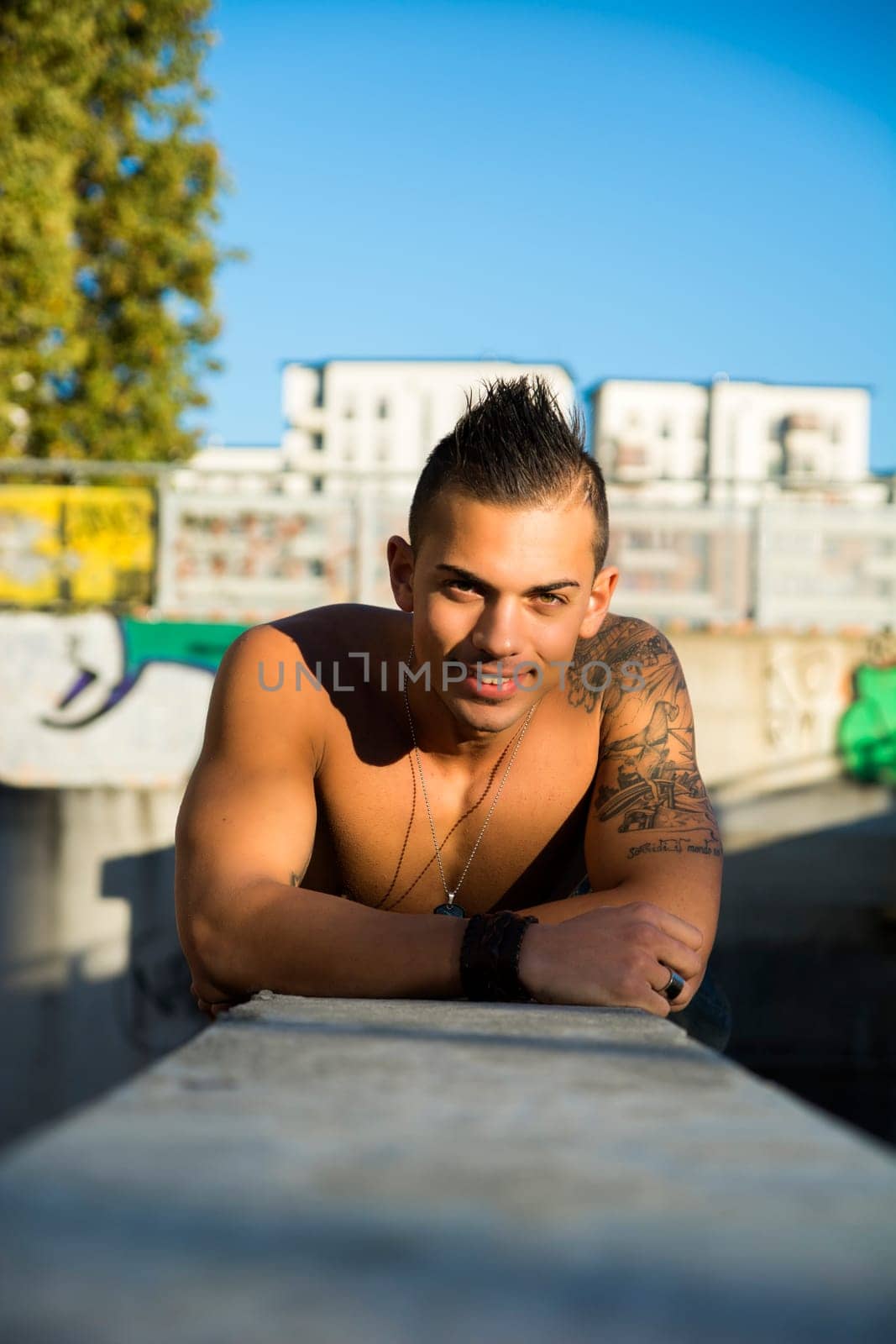 Handsome fit athletic shirtless young man in city setting leaning on concrete, looking at camera smiling