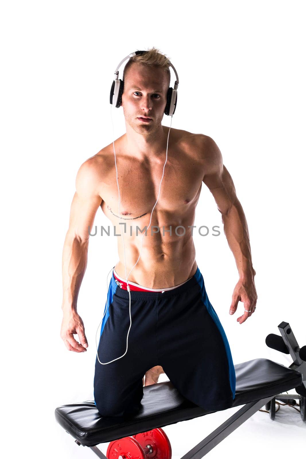 Muscular shirtless young man exercising with weights, bench, dumbbells, listening to music, isolated on white
