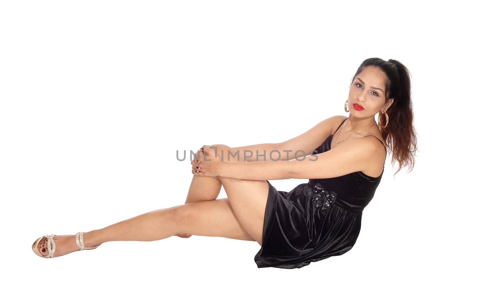 A beautiful woman in a short black dress sitting on the floor, holding.her leg and looking at the camera, isolated for white background.