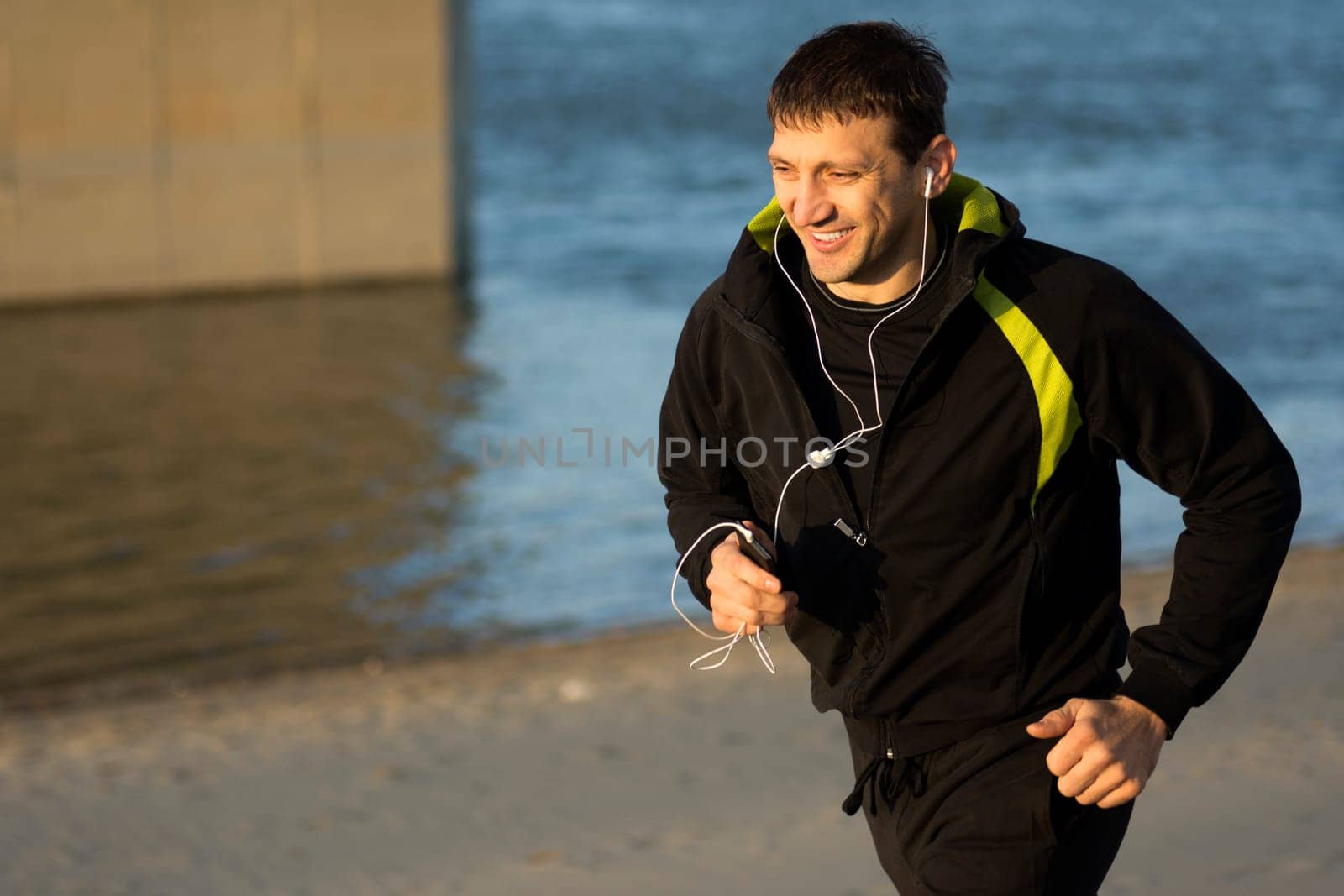 Smiling man is running with earphones outdoors on the beach.