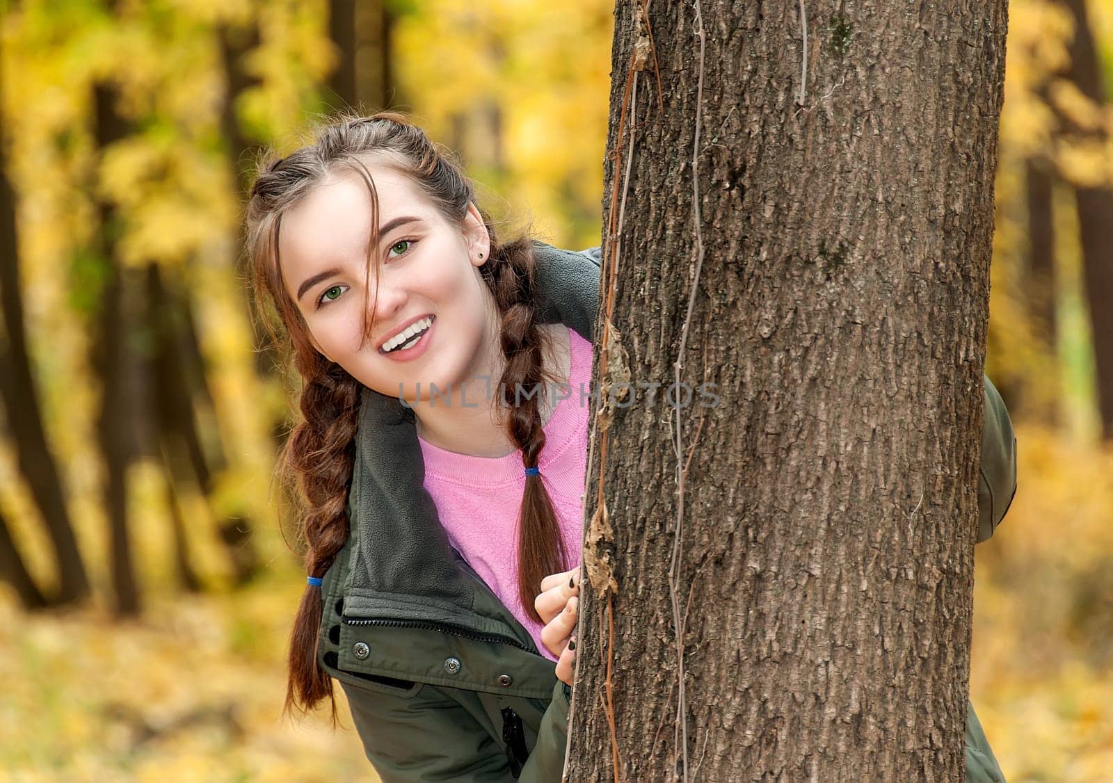 Laughing teen girl. A girl looks out from behind a tree in the park. Smiling happy face.
