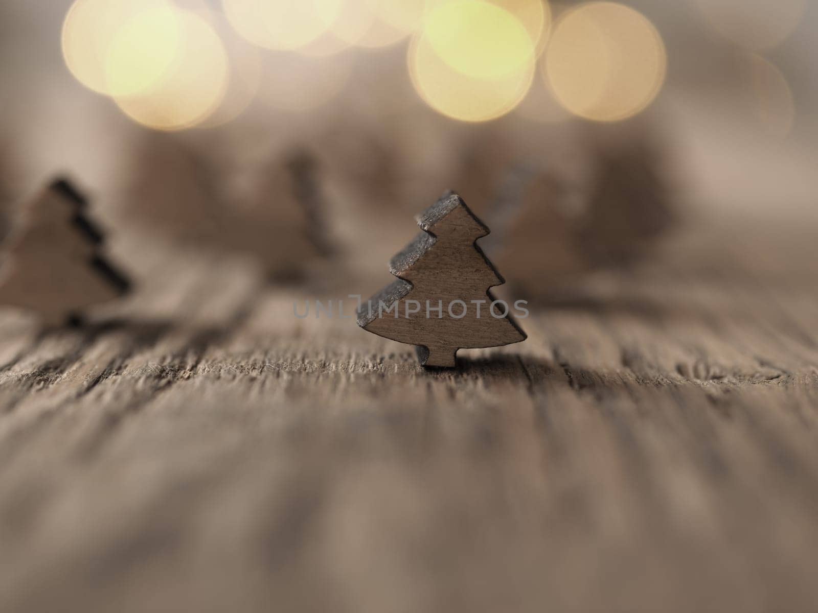 Little tree shapes on a wooden table with blurred Christmas lights in the background, selective focus on the first tree