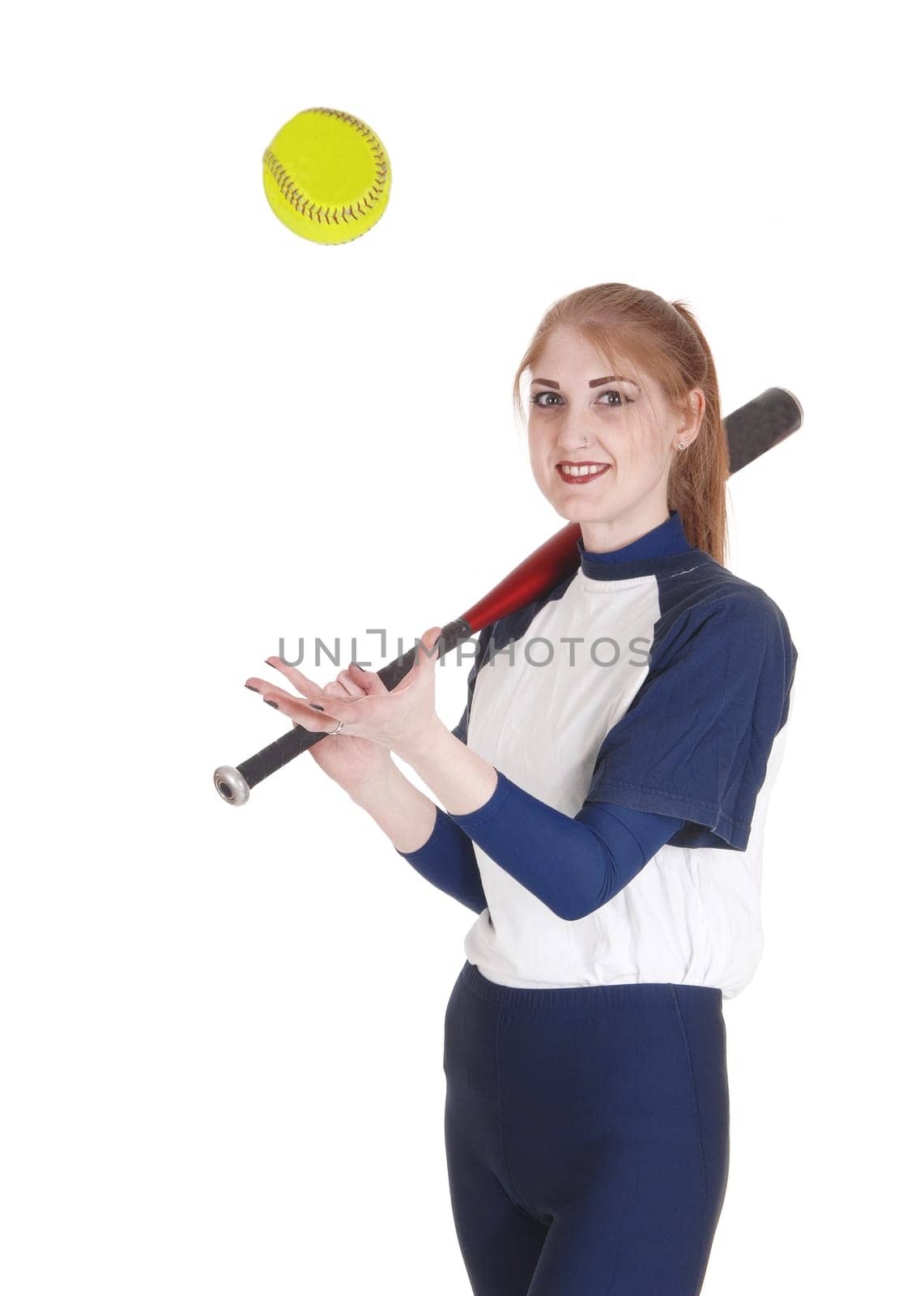 A smiling young woman standing in her blue softball uniform playing.with the yellow ball holding her bat, isolated for white background.